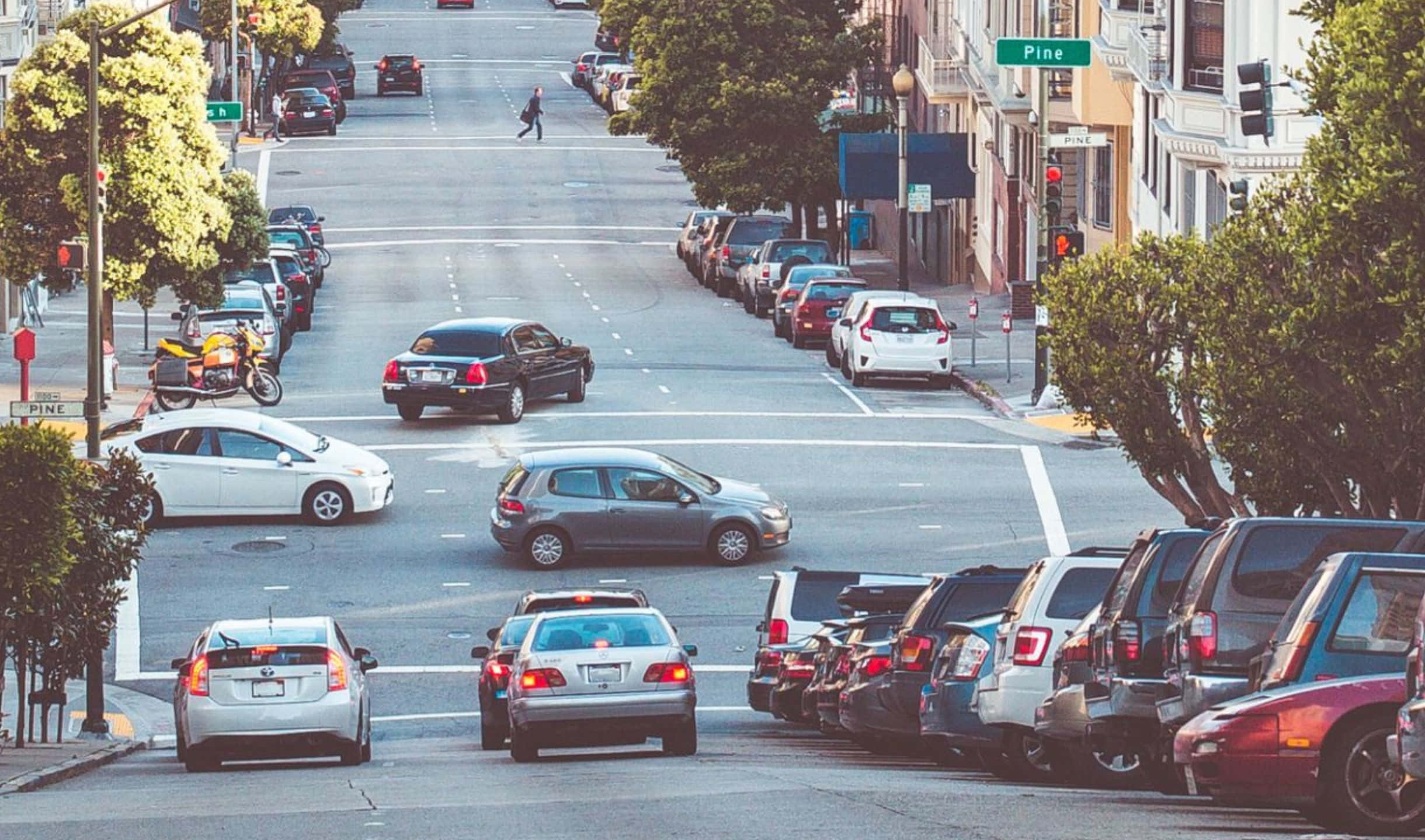 2440x1440 San Francisco 桌布 壁紙 — 一條繁忙的城市街道，兩旁停滿汽車，一名行人正在明亮的日光下過馬路。