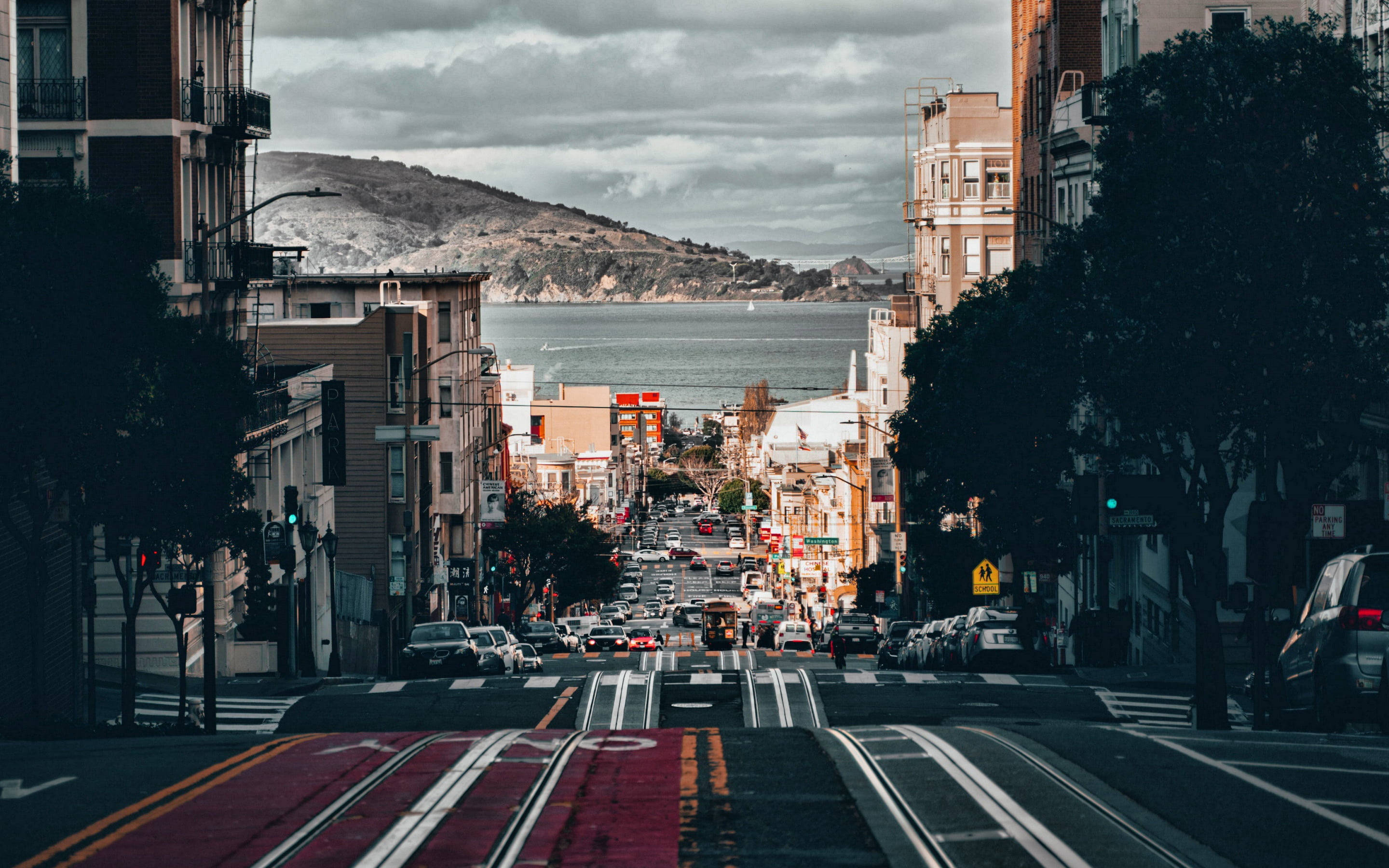 San Francisco Phone 桌布 壁紙 — 一條繁忙的城市街道，有纜車軌道，通向海灣和遠處的山丘，天空多雲。
