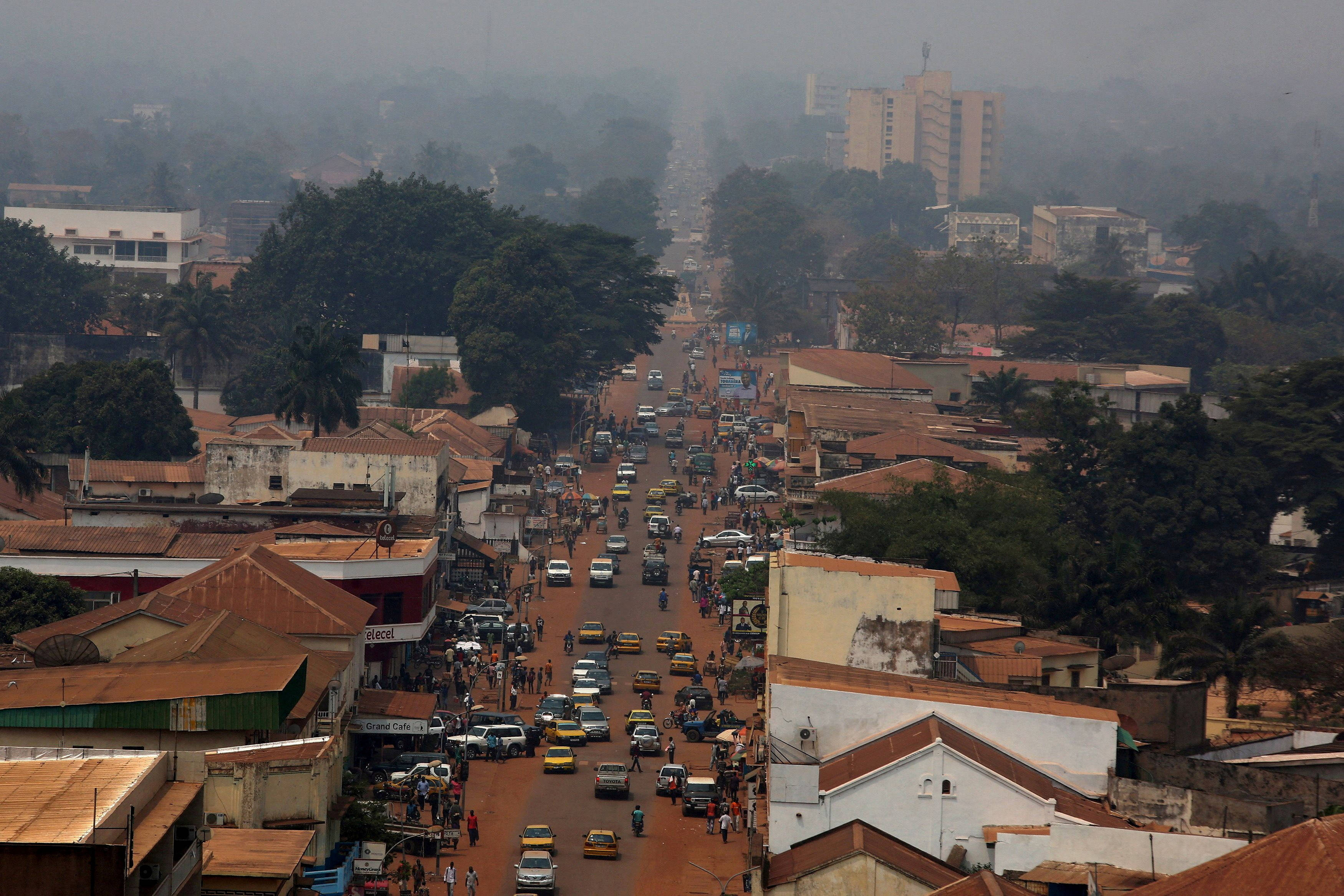 城市 桌布 壁紙 — 一條繁忙的城市街道，汽車和行人穿梭其中，蜿蜒穿過霧氣瀰漫的城市景觀，混合建築風格和茂密的綠色樹木。