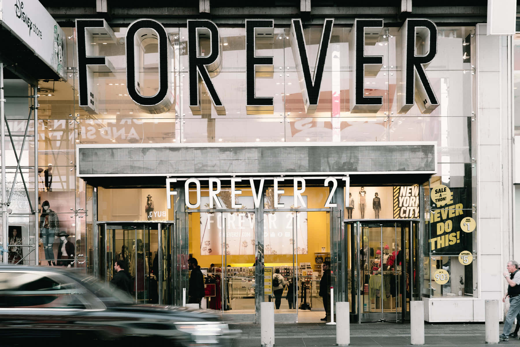 永遠21 桌布 壁紙 — 一條繁忙的城市街道上有一間Forever 21商店，設有大型玻璃窗和旋轉門，行人及一輛模糊的汽車經過。