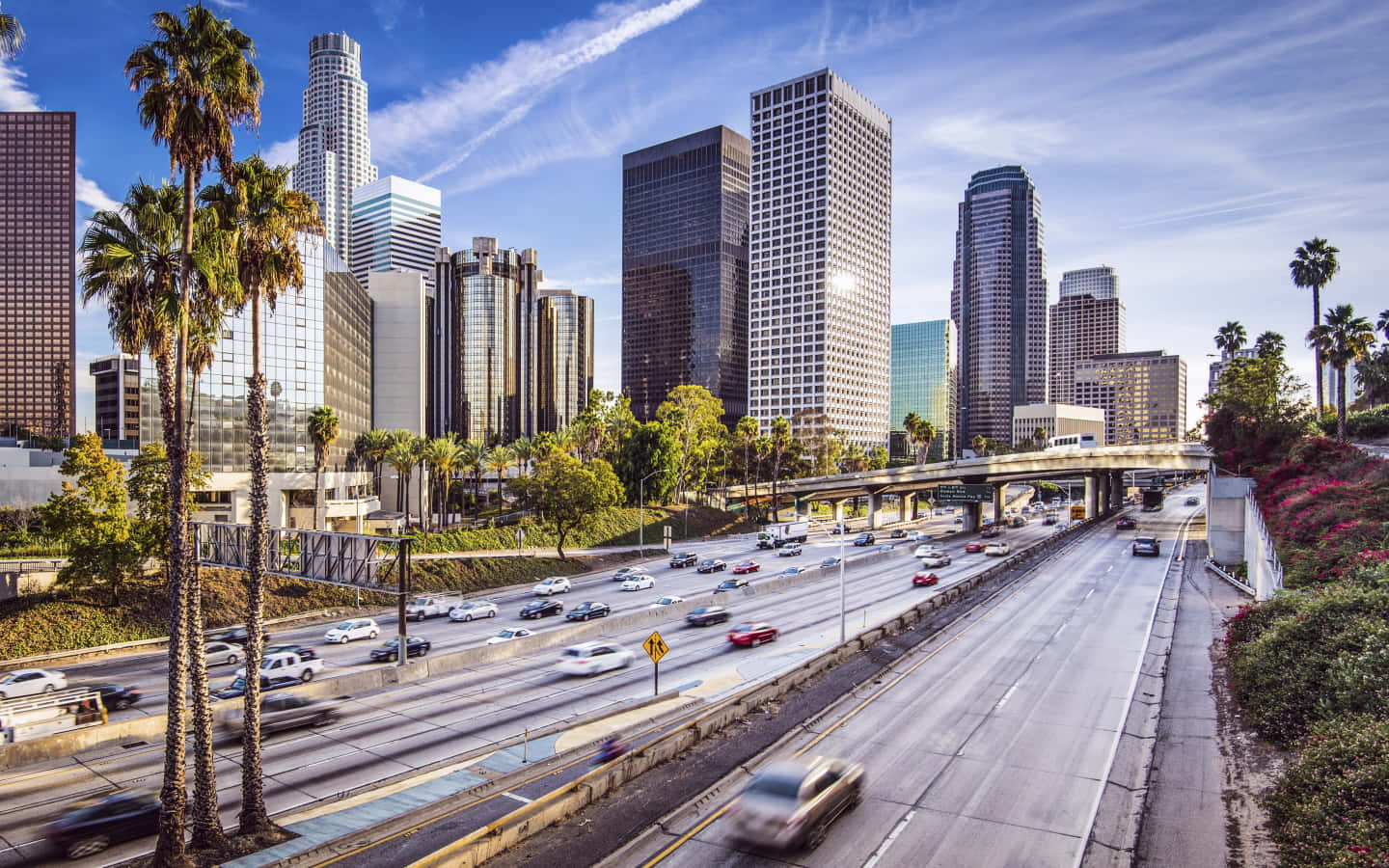 La 桌布 壁紙 — 一條繁忙的城市高速公路，兩旁種有棕櫚樹和高聳的摩天大樓，天空湛藍，白雲輕飄。
