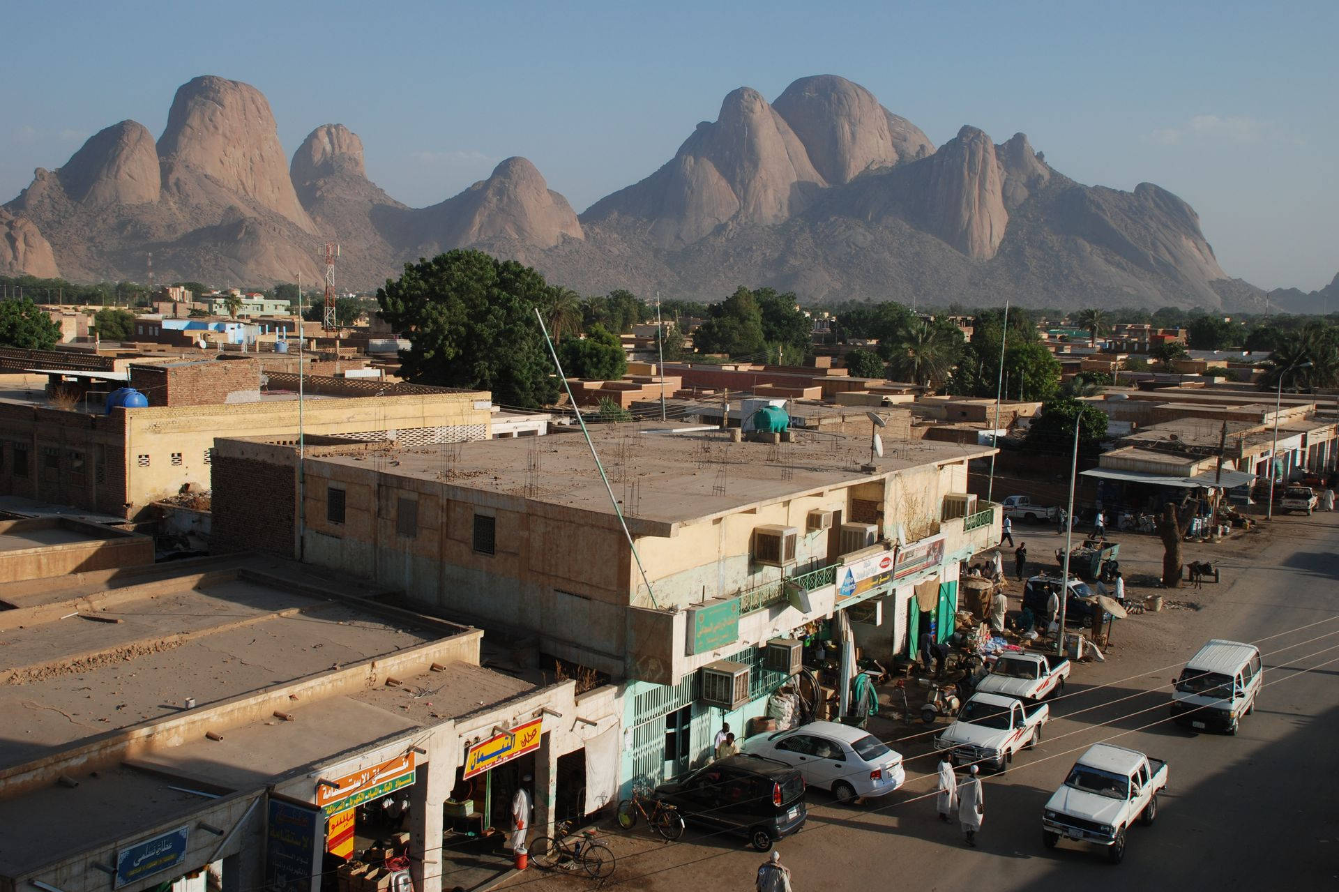Sudan 桌布 壁紙 — 一條繁忙的城鎮街道，低矮建築物和汽車在晴朗的天空下，遠方聳立的岩石山脈框住景色。