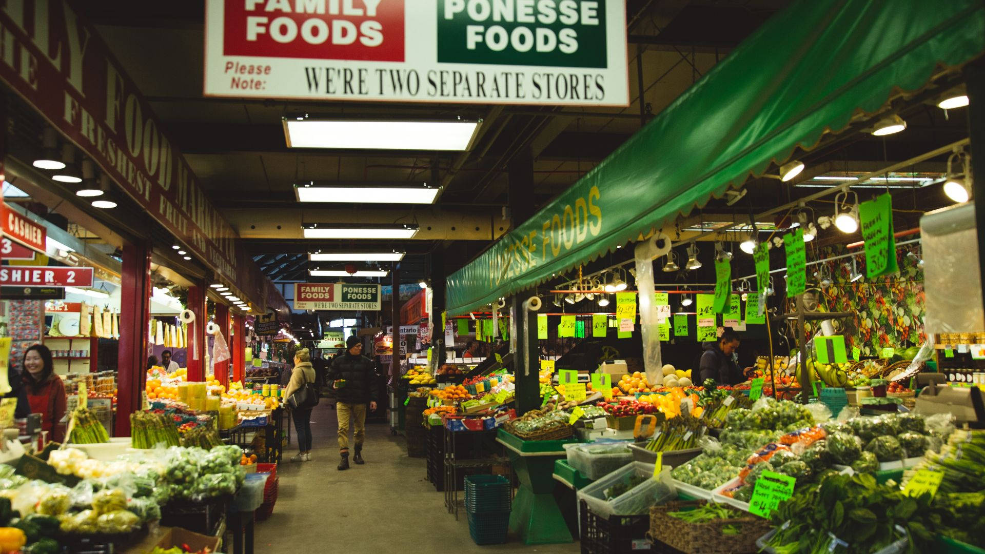 Vegetables 桌布 壁紙 — 一條繁忙的室內市場通道，兩旁擺滿色彩繽紛的農產品攤位，上方有綠色遮陽篷和明亮的螢光燈。