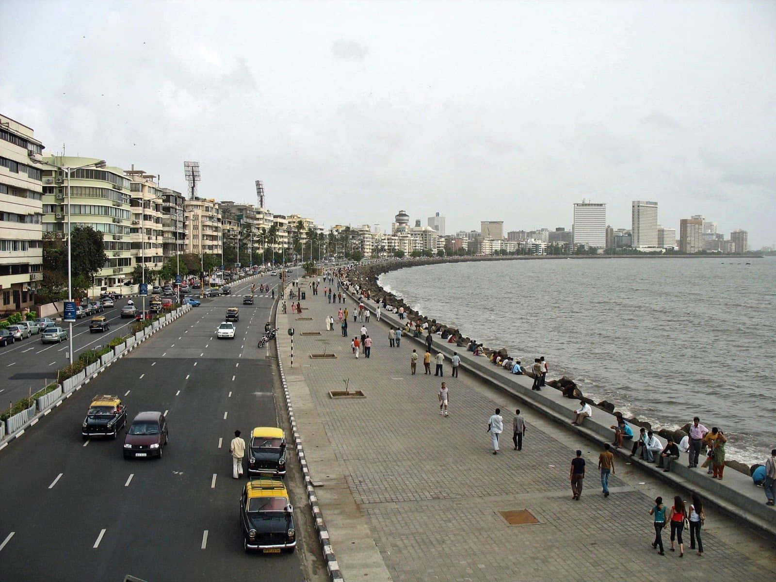 Mumbai 桌布 壁紙 — 一條繁忙的海濱長廊，行人與黃色計程車沿著海邊的寬闊道路行駛，天空多雲。