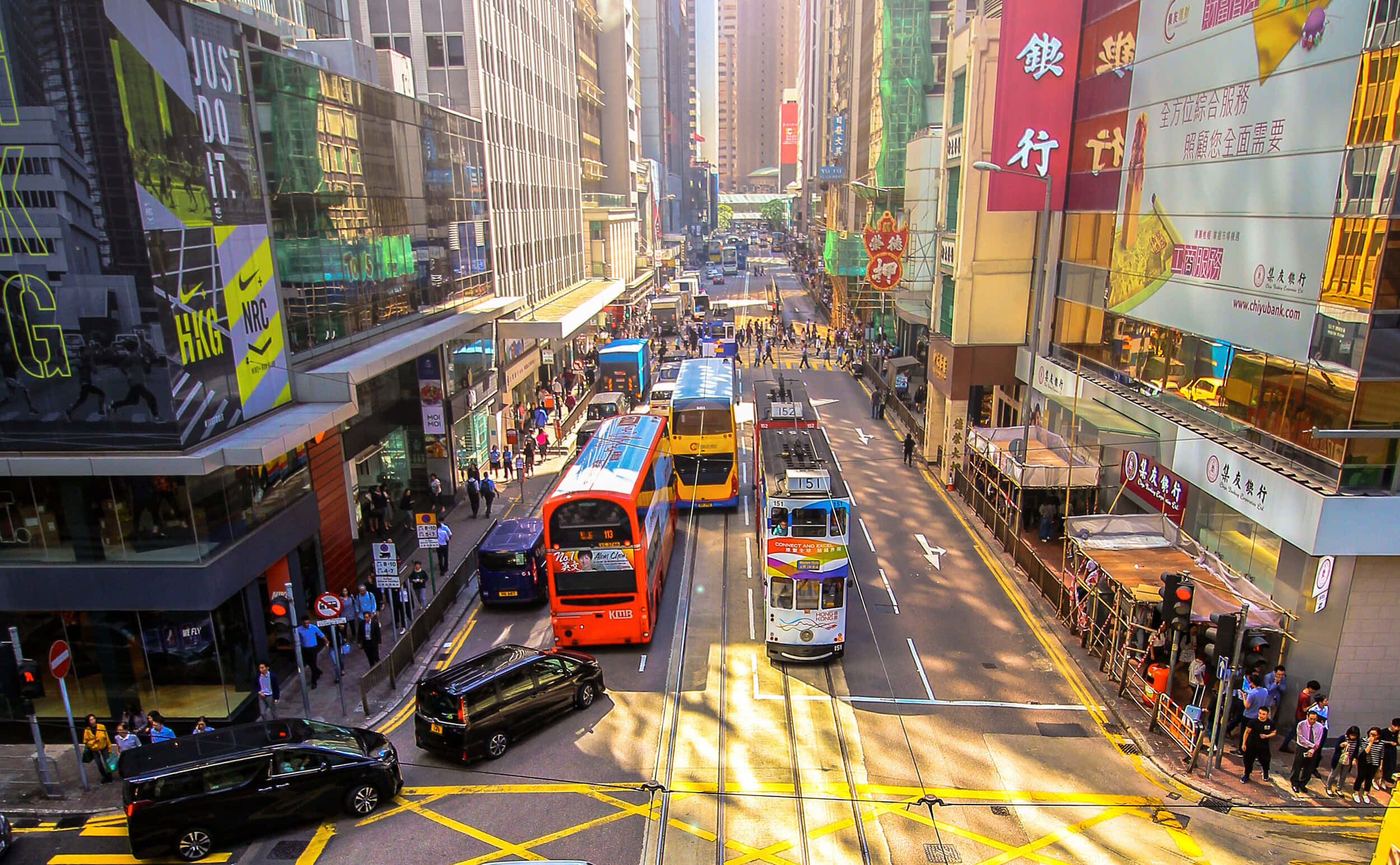 香港 桌布 壁紙 — 一條繁忙的香港街道，充滿色彩繽紛的雙層巴士、電車和行人，在明亮的陽光下投下長長的影子。