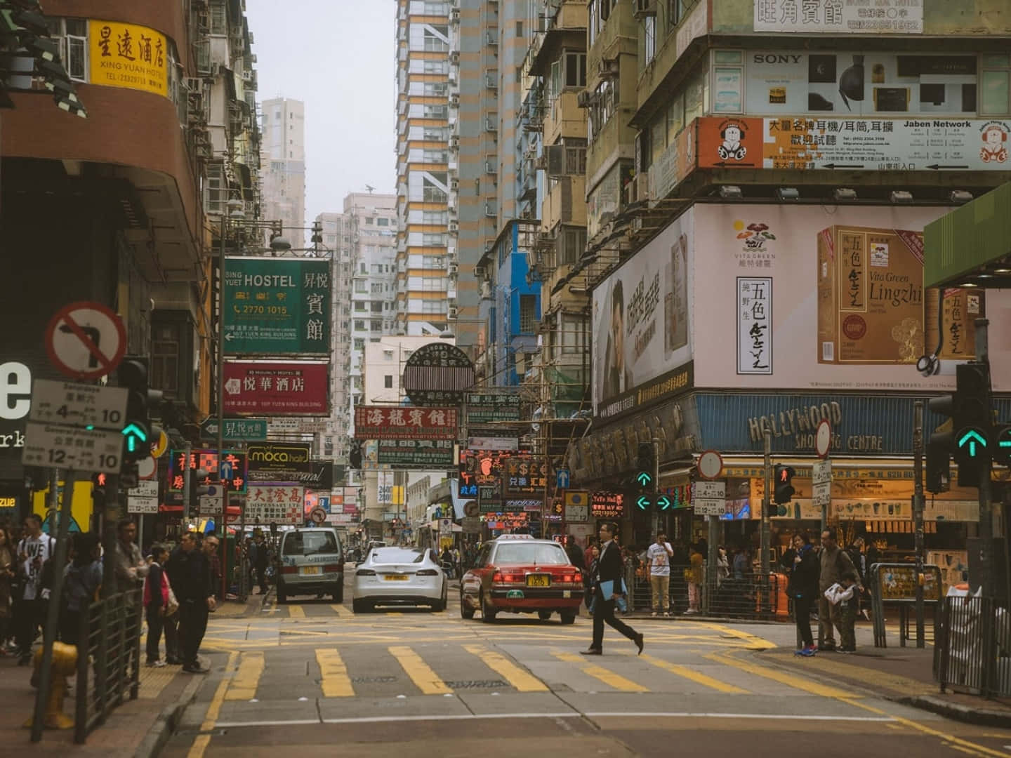 香港 桌布 壁紙 — 一條繁忙的香港街道，擠滿行人、汽車，以及裝飾著鮮豔霓虹招牌和密集廣告的高聳建築物。