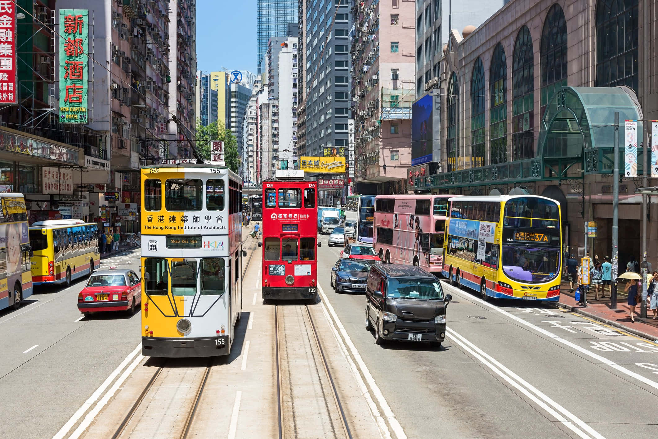香港 桌布 壁紙 — 一條繁忙的香港街道，標誌性的紅色和黃色電車與巴士和汽車在明亮的藍天下共用車道。