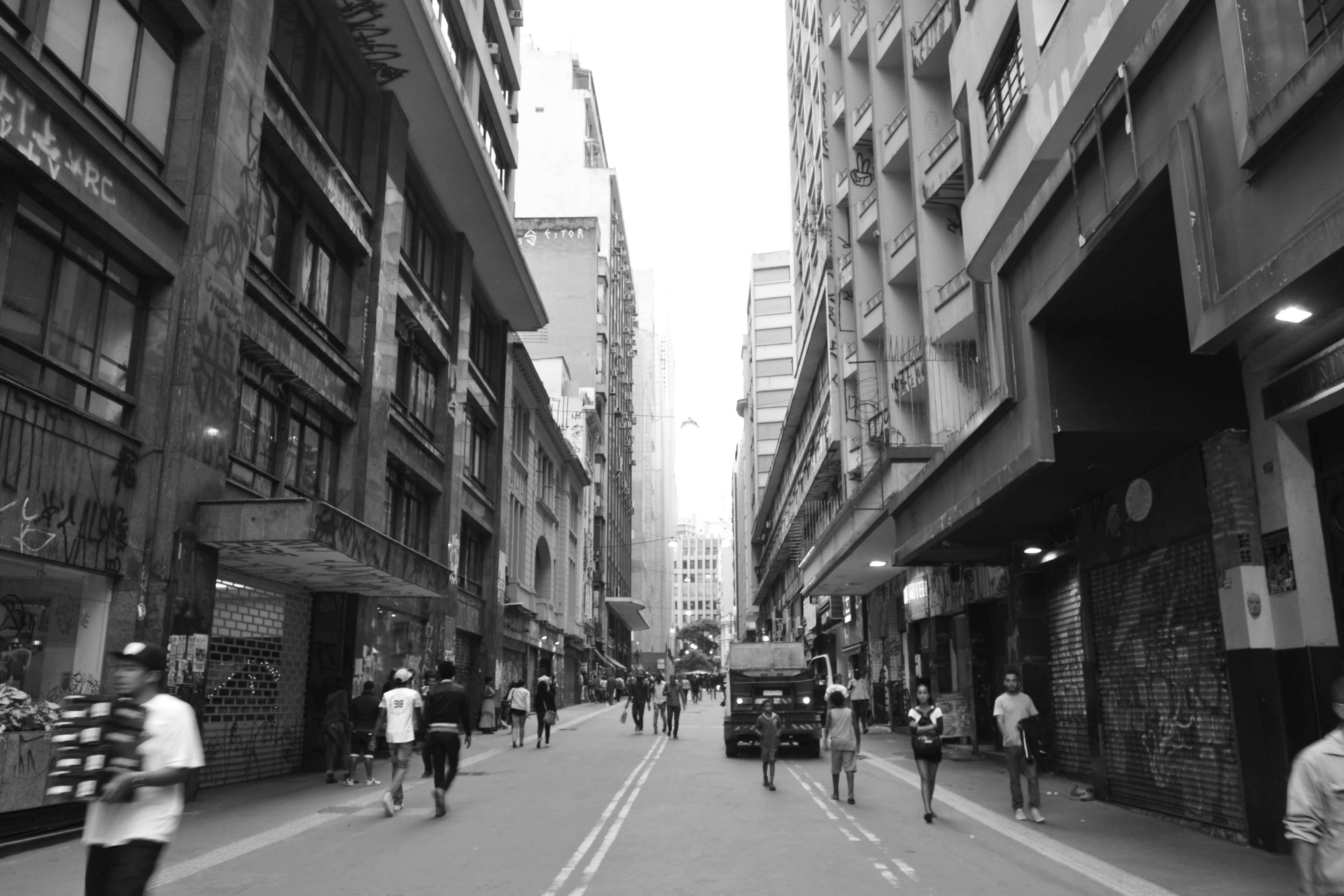 聖保羅 桌布 壁紙 — 一條繁忙的黑白城市街道，行人和一輛卡車在有塗鴉的高樓之間穿梭。
