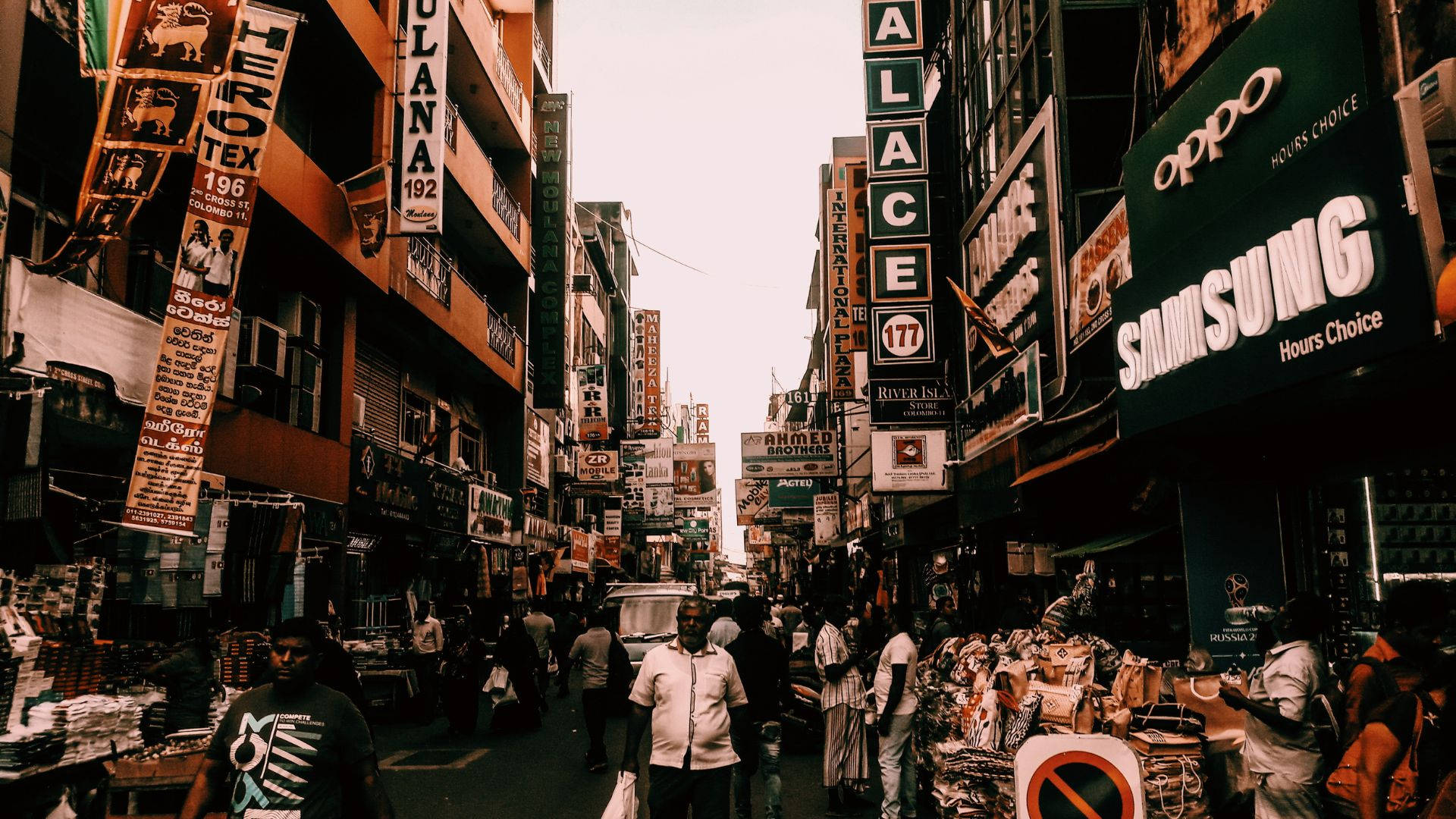 crowd 桌布 壁紙 — 一條繁華的城市小巷，擠滿了行人和滿溢的市場攤位，頭頂上掛滿招牌和商店門面。