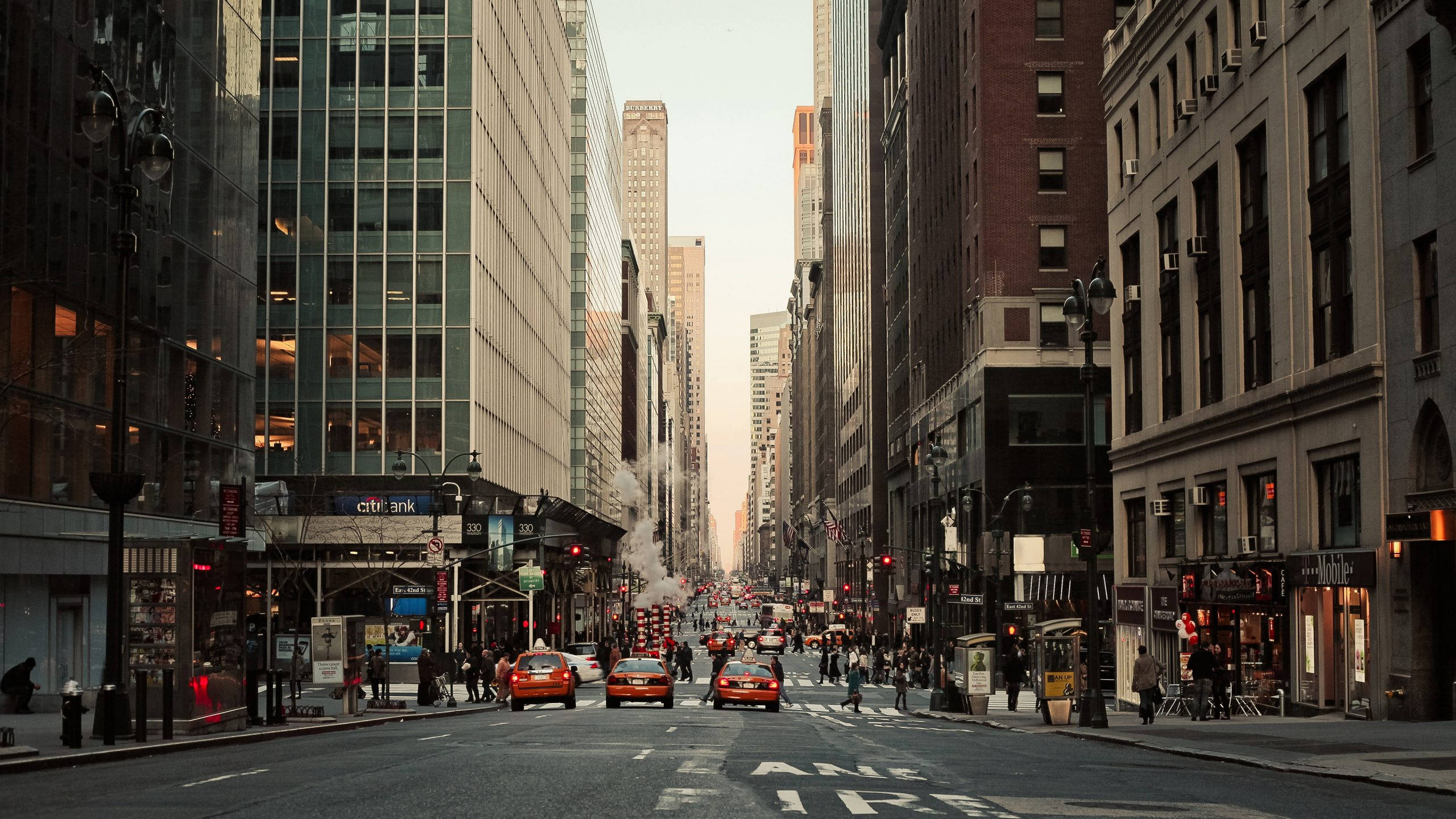 New York Street 桌布 壁紙 — 一條繁華的城市街道，兩旁高聳的摩天大樓和紅色計程車，籠罩在朦朧的金色夕陽下。