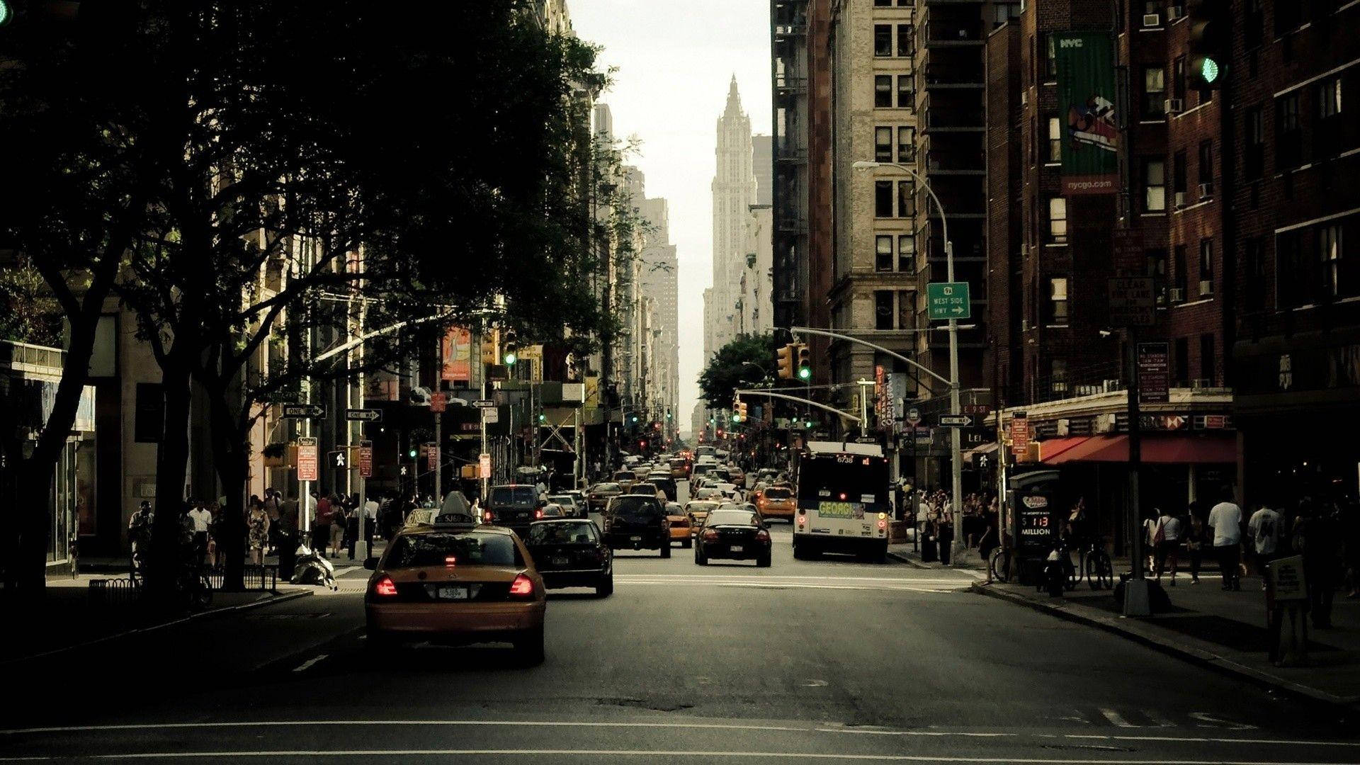 New York 桌布 壁紙 — 一條繁華的紐約市街道，兩旁高樓大廈和樹木林立，在朦朧的天空下擠滿了黃色的士和行人。