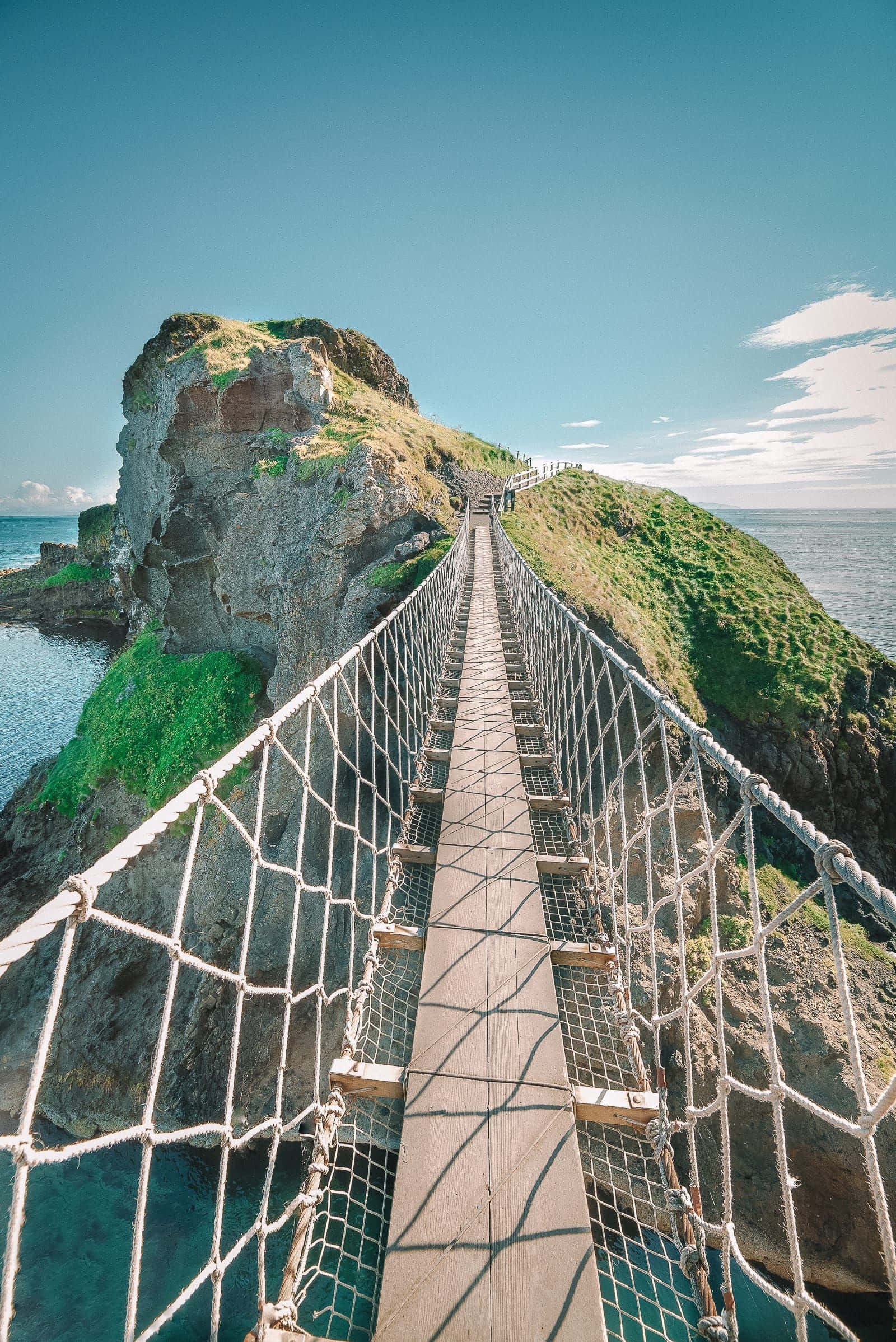 bridge 桌布 壁紙 — 一條繩索吊橋橫跨碧綠色的海水，連接到一片草地懸崖，上方是清澈的藍天。