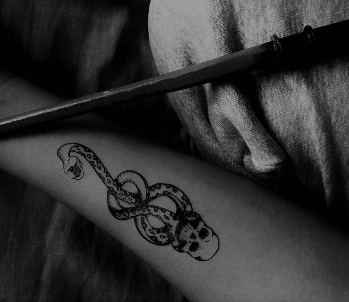 Death Eaters 桌布 壁紙 — 一條纏繞著骷髏頭的蛇紋身手臂黑白特寫，旁邊放著一把刀。
