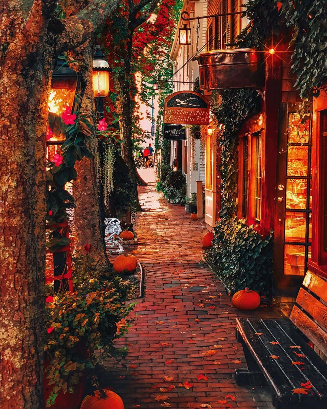 Fall Town 桌布 壁紙 — 一條舒適的秋季小巷，兩旁是磚砌建築、常春藤、南瓜和發光的燈籠，沐浴在溫暖的金色光芒中。