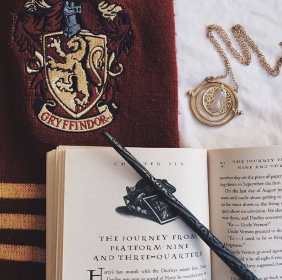 hp aesthetic 桌布 壁紙 — 一條葛萊芬多圍巾、一個時間轉換器和一根魔杖放在白色表面上的一本打開的哈利波特書上。
