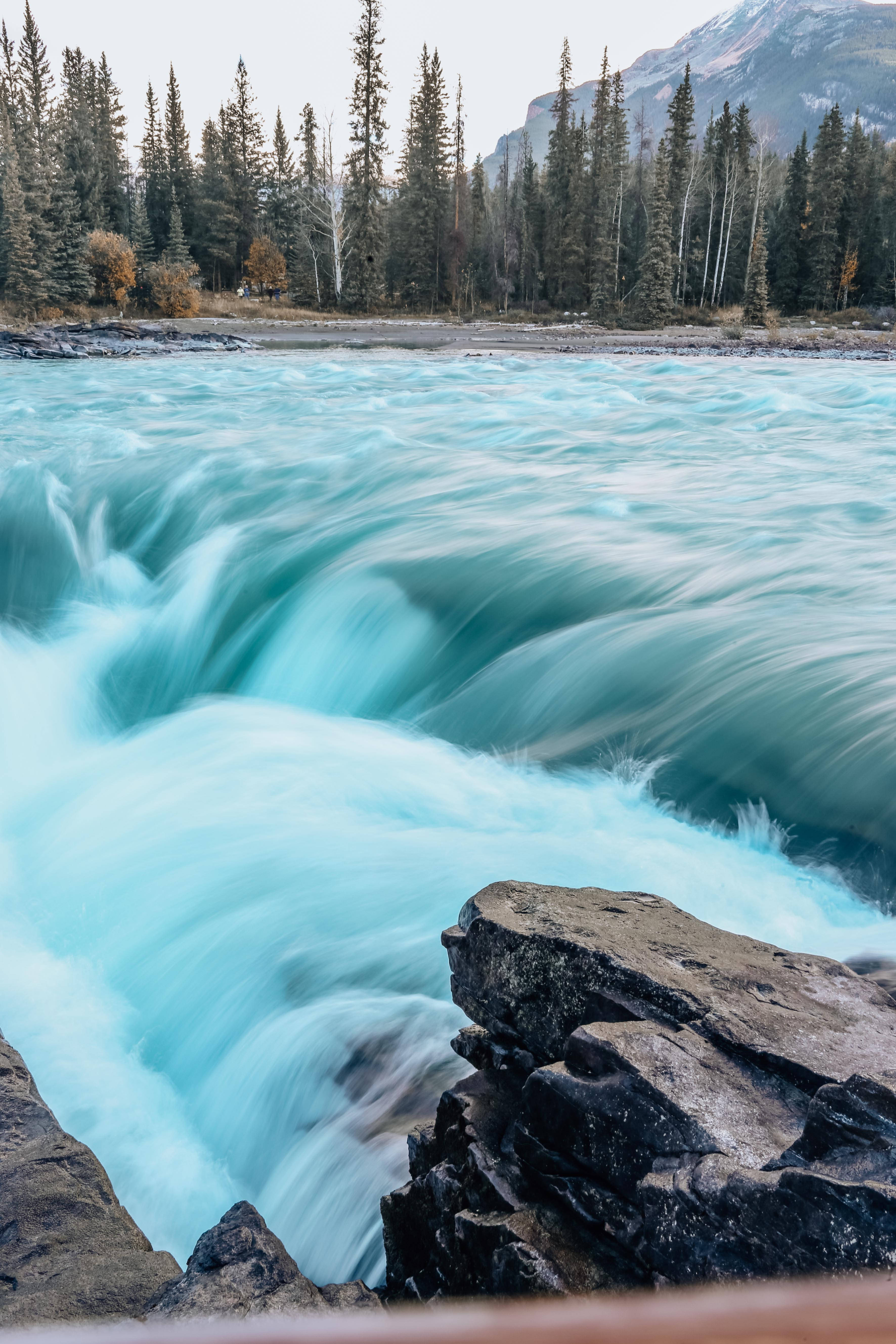 Falling Water 桌布 壁紙 — 一條藍綠色的河流衝過岩石，帶有霧狀動態模糊，由常綠樹和遠山框住。