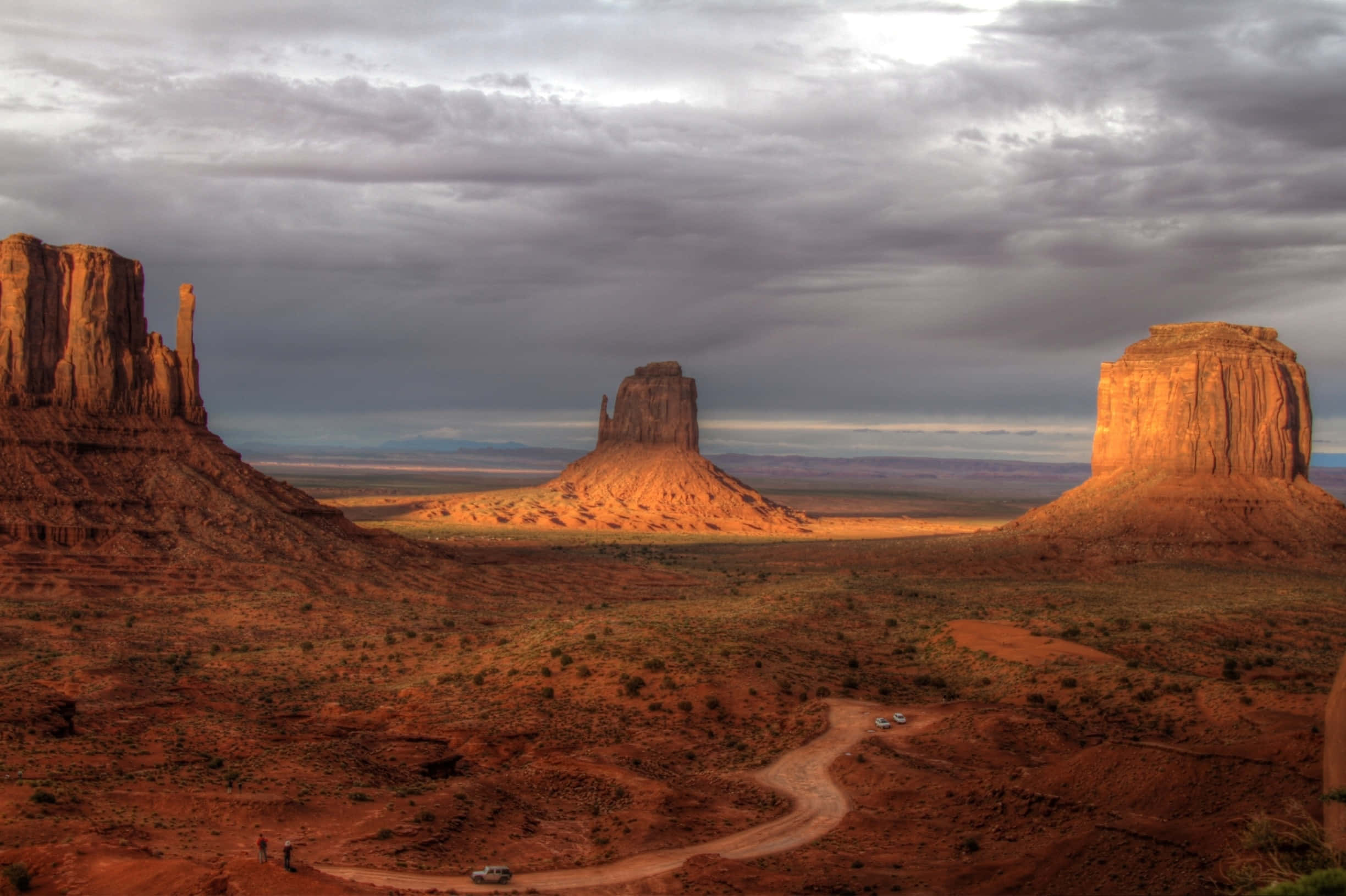 Arizona 桌布 壁紙 — 一條蜿蜒的土路穿過廣闊的沙漠景觀，在戲劇性的多雲天空下，聳立著紅色的岩石山丘。