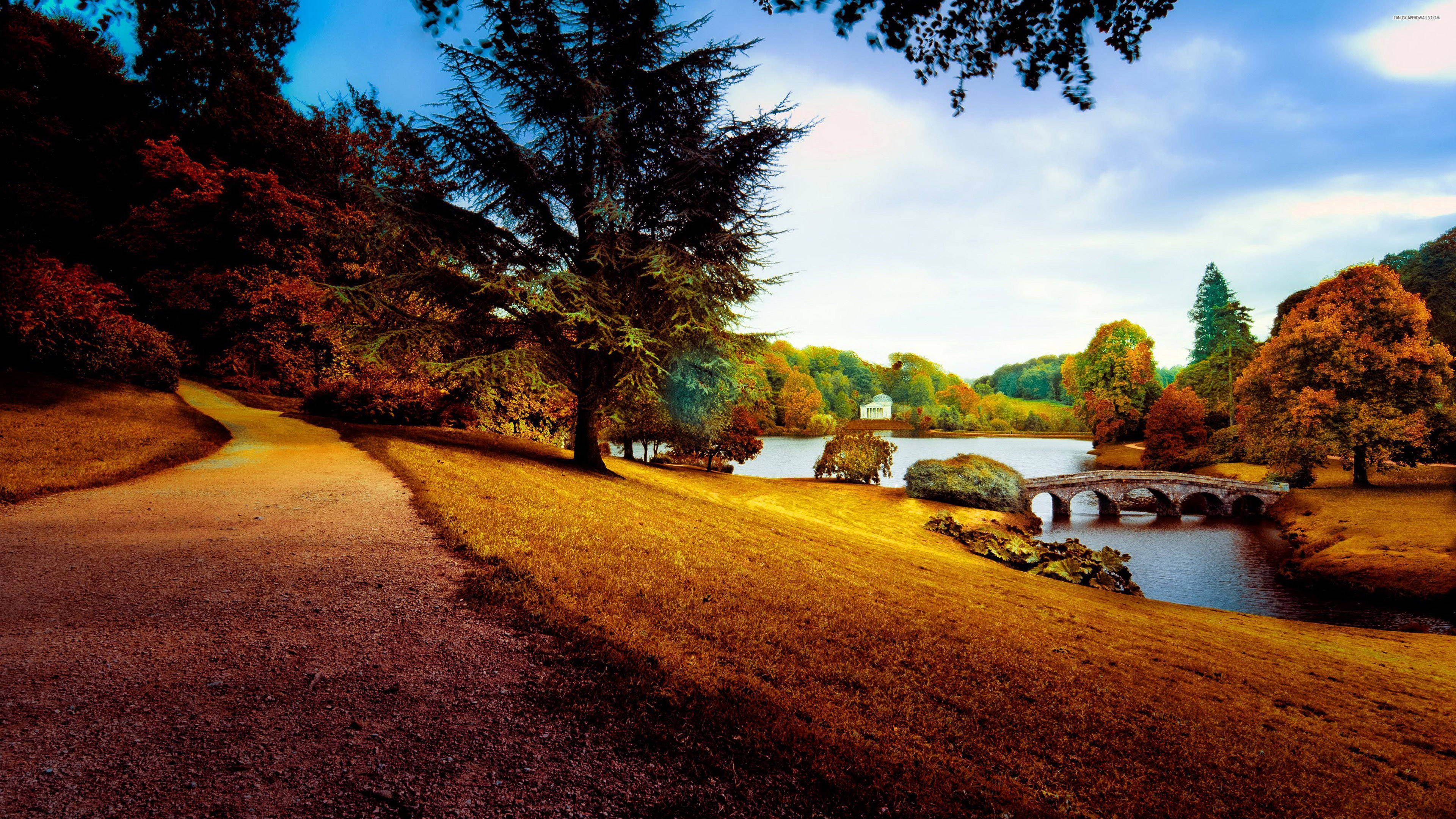Autumn Season 桌布 壁紙 — 一條蜿蜒的小路通往一座石橋，橫跨在平靜的湖面上，周圍是秋天的樹木，天空柔和湛藍。