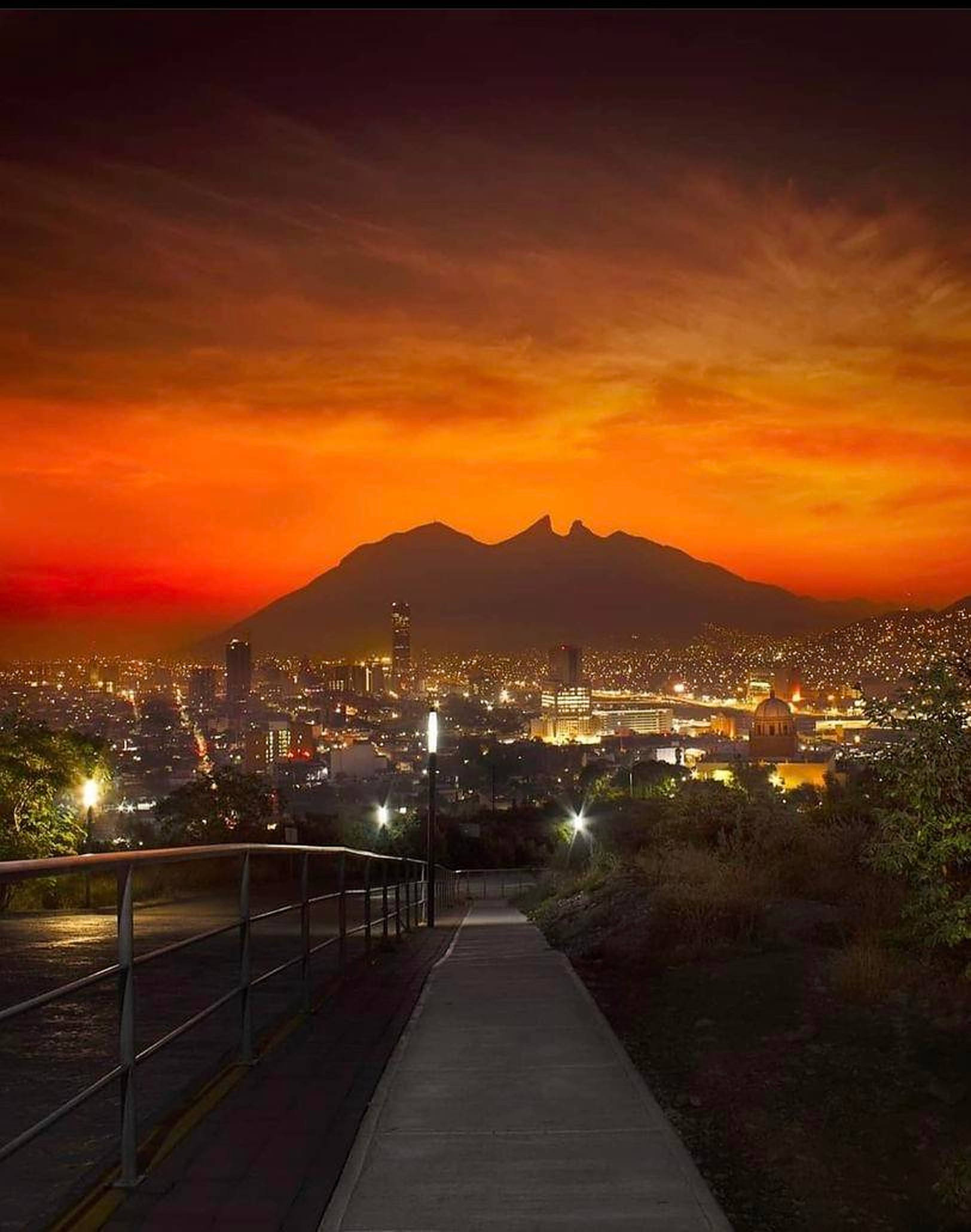 Monterrey 桌布 壁紙 — 一條蜿蜒的小路通往日落時分閃閃發光的城市景觀，黑暗的山脈輪廓映襯著火紅的橙色雲彩。
