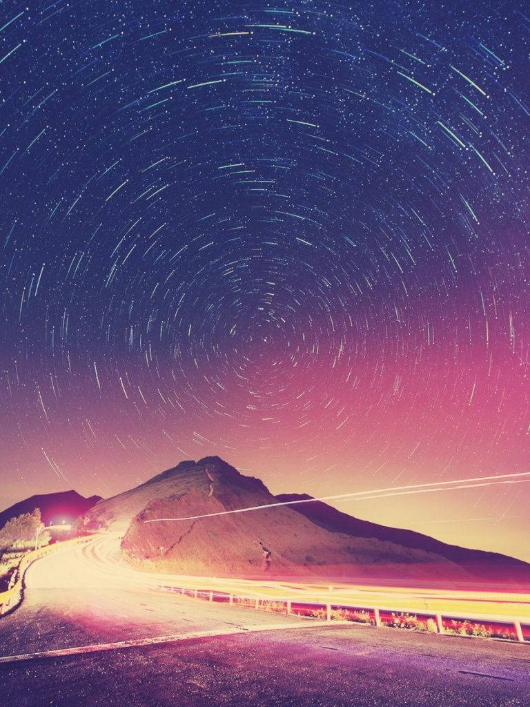 在帶有星軌和粉紫色調的天空下蜿蜒的山路。 — iPad mini 動態圖片 壁紙