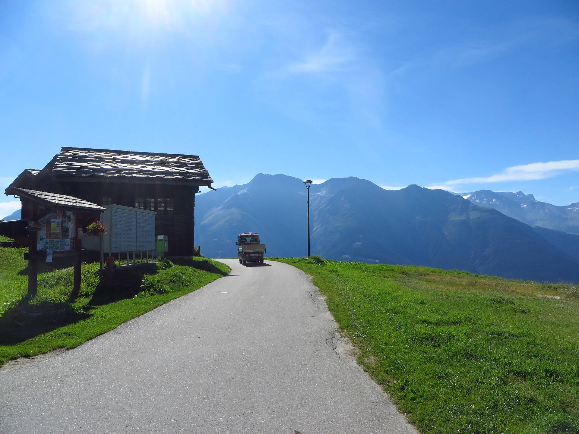 Alps 桌布 壁紙 — 一條蜿蜒的山路在明亮的藍天下繞過一間木屋，遠處有白雪覆蓋的山峰。