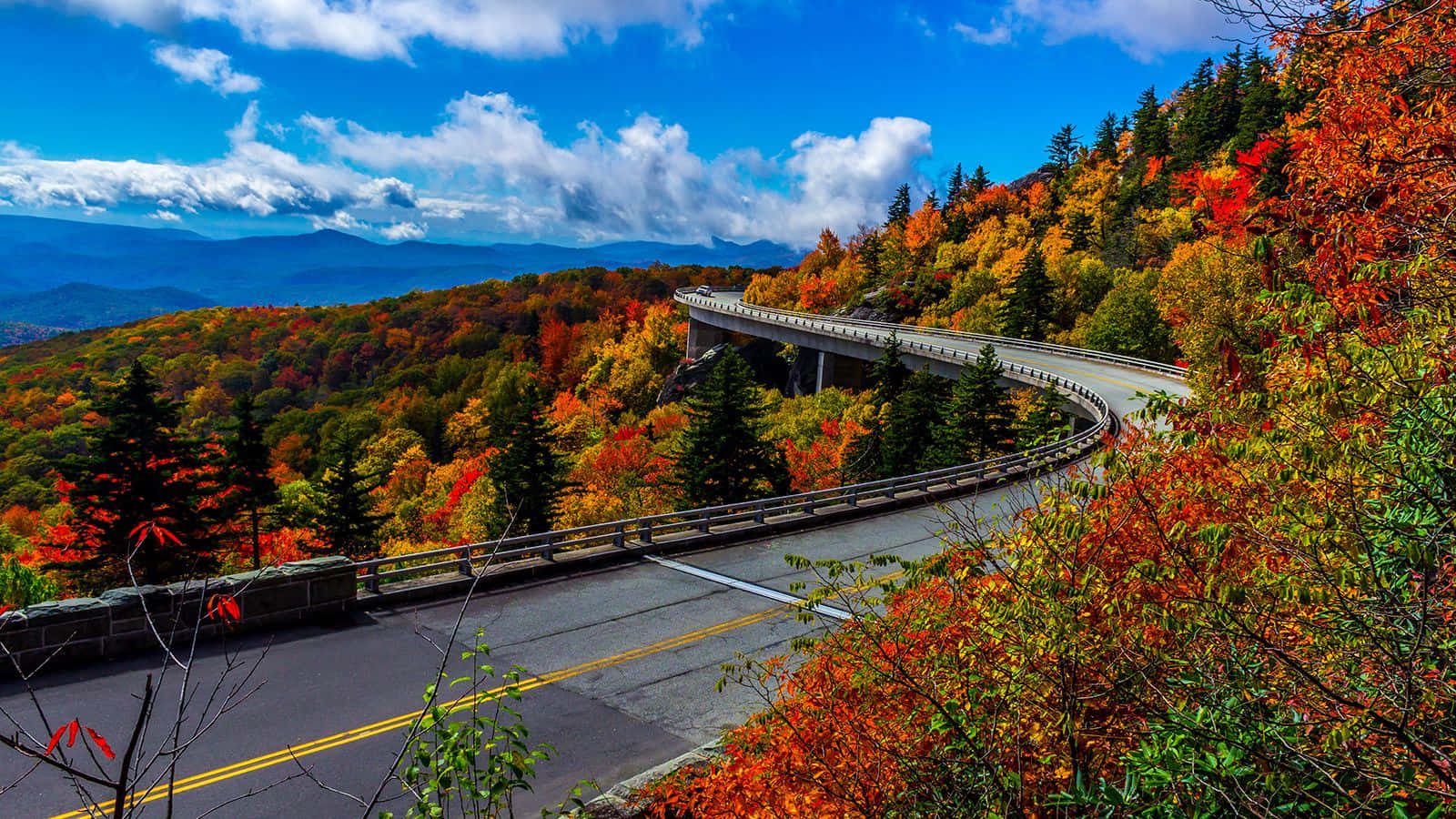 Fall Road 桌布 壁紙 — 一條蜿蜒的山路在明亮的藍天白雲下穿過充滿活力的秋葉。