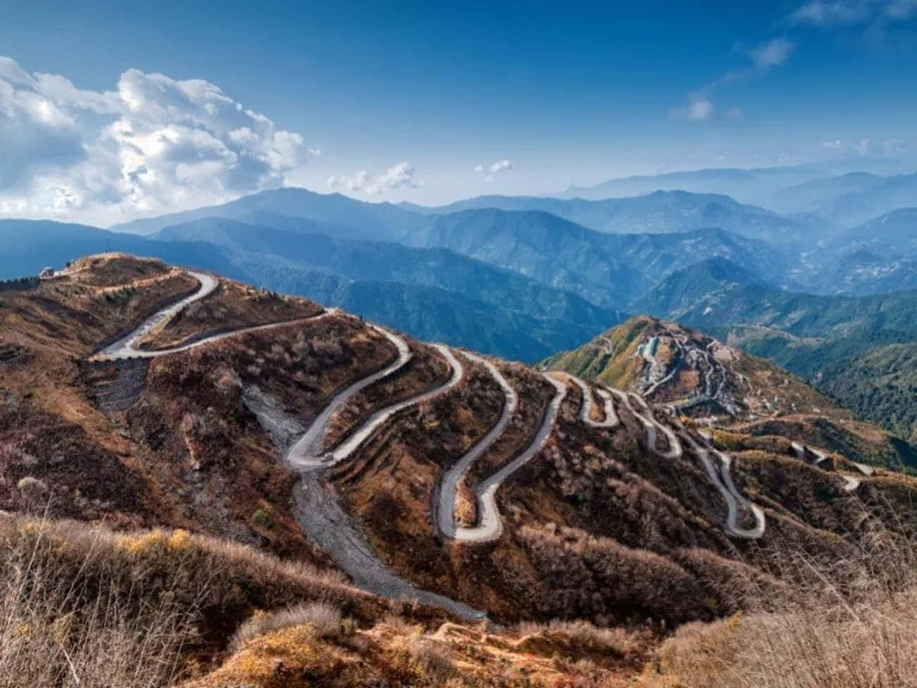 風景 桌布 壁紙 — 一條蜿蜒的山路在明亮的藍天白雲下蜿蜒穿過秋日山丘。