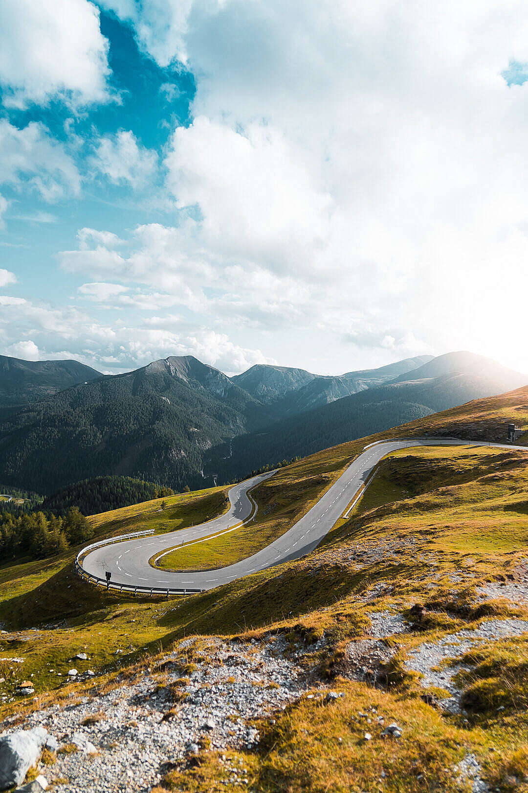 Austria 桌布 壁紙 — 一條蜿蜒的山路在陽光明媚的綠色山丘中蜿蜒而行，頭頂是明亮的藍天和蓬鬆的白雲。