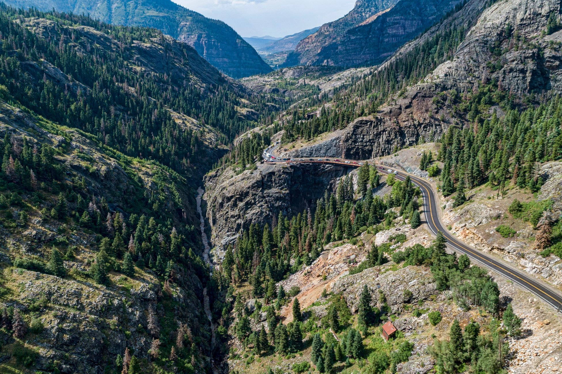 Colorado 桌布 壁紙 — 一條蜿蜒的山路穿過一個深林峽谷，兩旁是陡峭的岩石懸崖，遠處有一座橋樑，天空明亮。