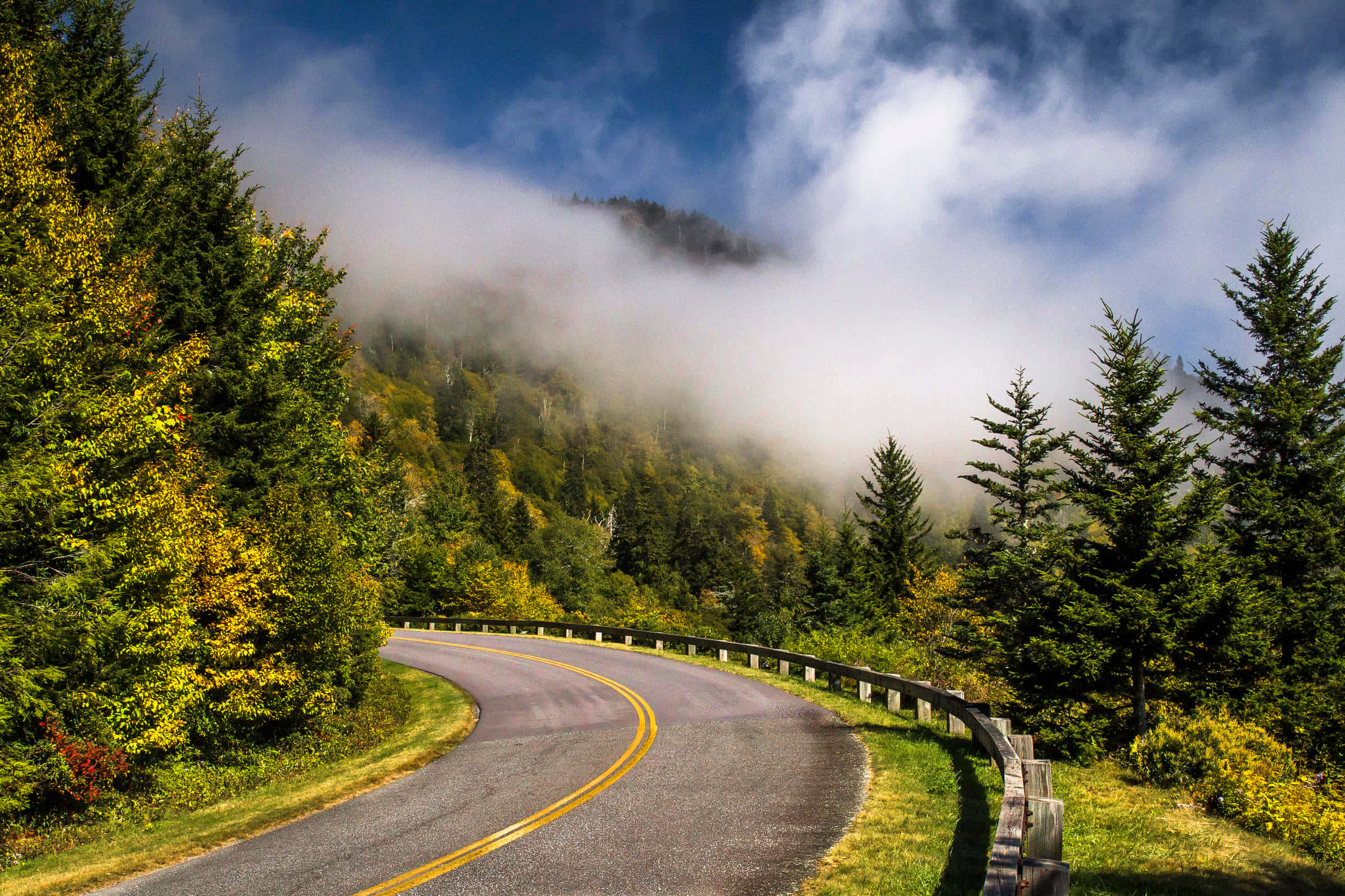 Blue Ridge Parkway 桌布 壁紙 — 一條蜿蜒的山路穿過秋天的森林，霧氣依附在山峰上，天空湛藍明亮。