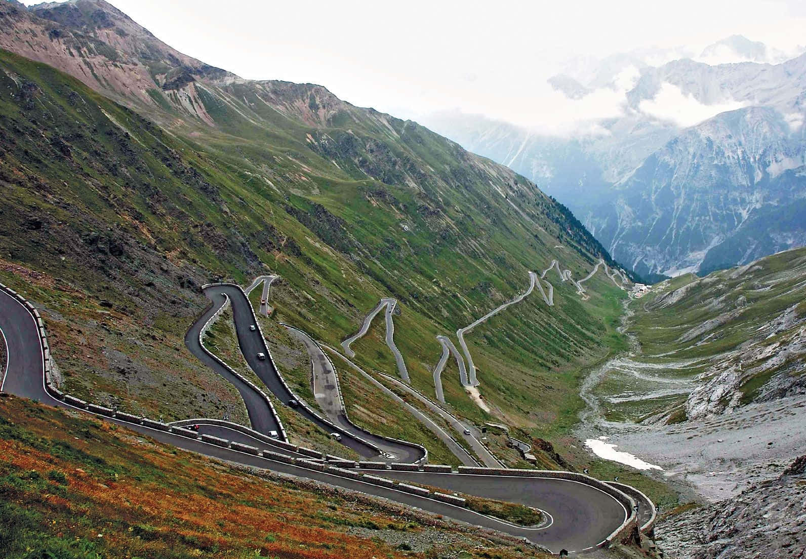 Road 桌布 壁紙 — 一條蜿蜒的山路穿過綠色和岩石斜坡，遠處有白雪覆蓋的山峰，天空朦朧。