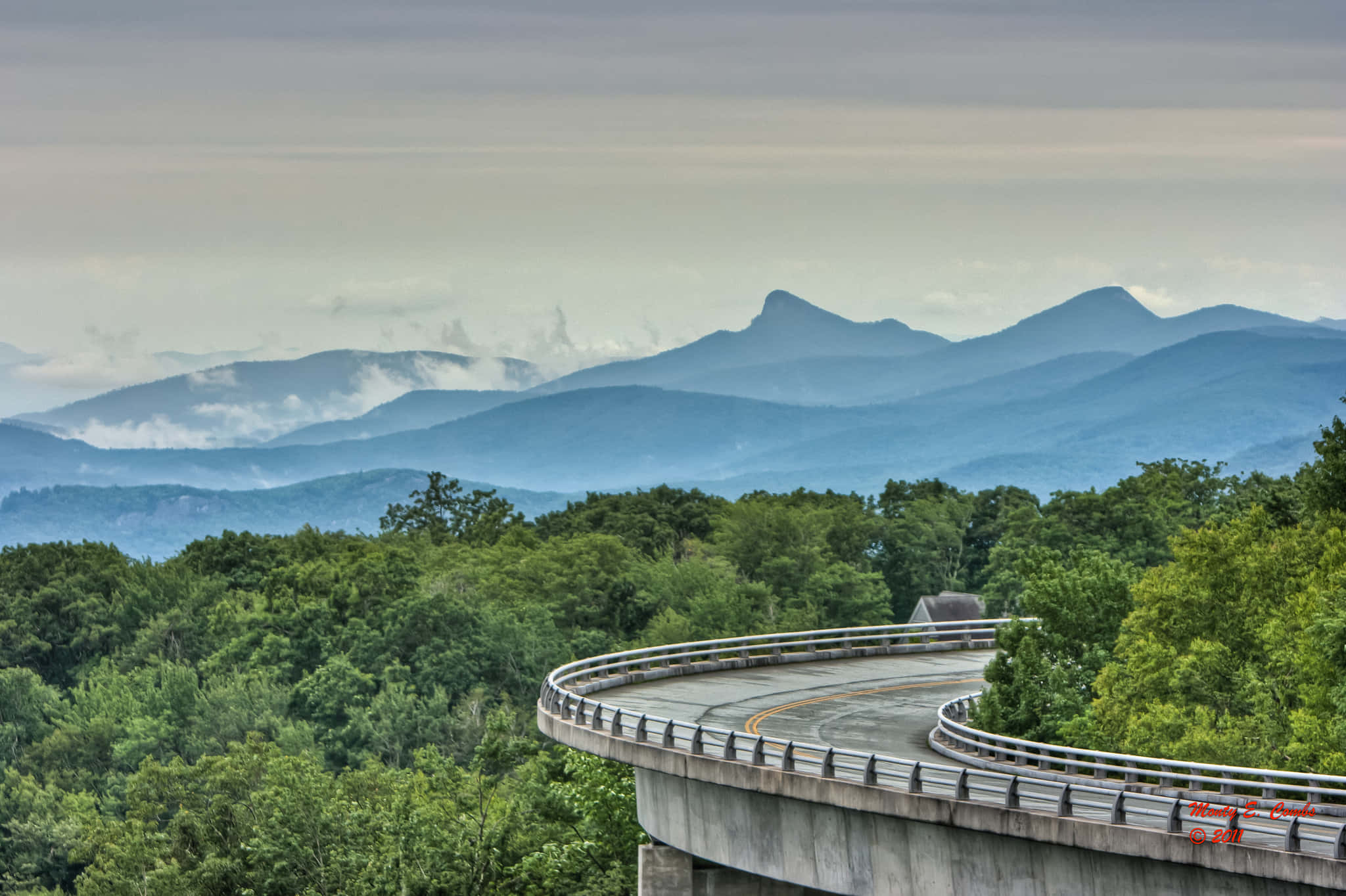 Blue Ridge Parkway 桌布 壁紙 — 一條蜿蜒的山路穿過茂密的綠色森林，遠處霧藍色的山峰在陰天中升起。