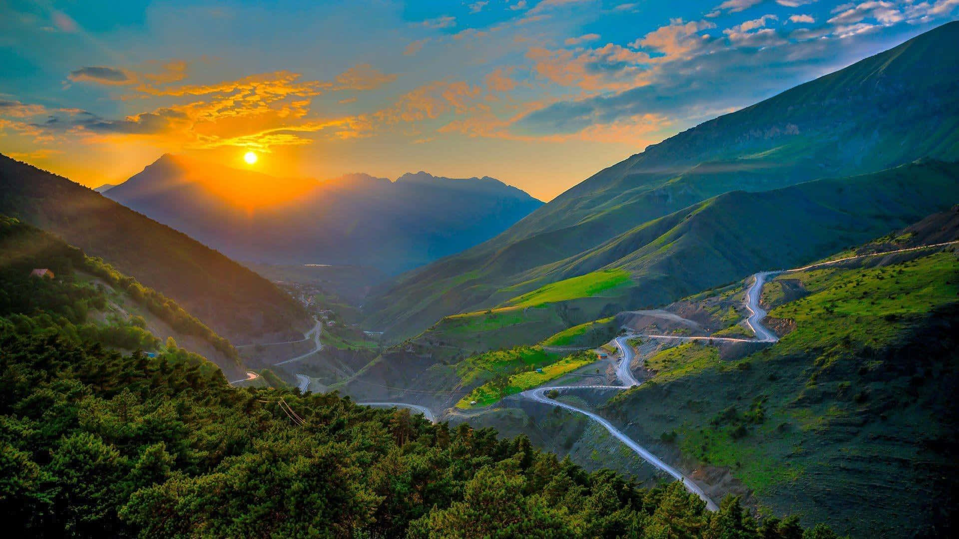 Beautiful Mountain 桌布 壁紙 — 一條蜿蜒的山路穿過鬱蔥的綠色山谷，夕陽在遠山背後落下，灑下金色的光芒。