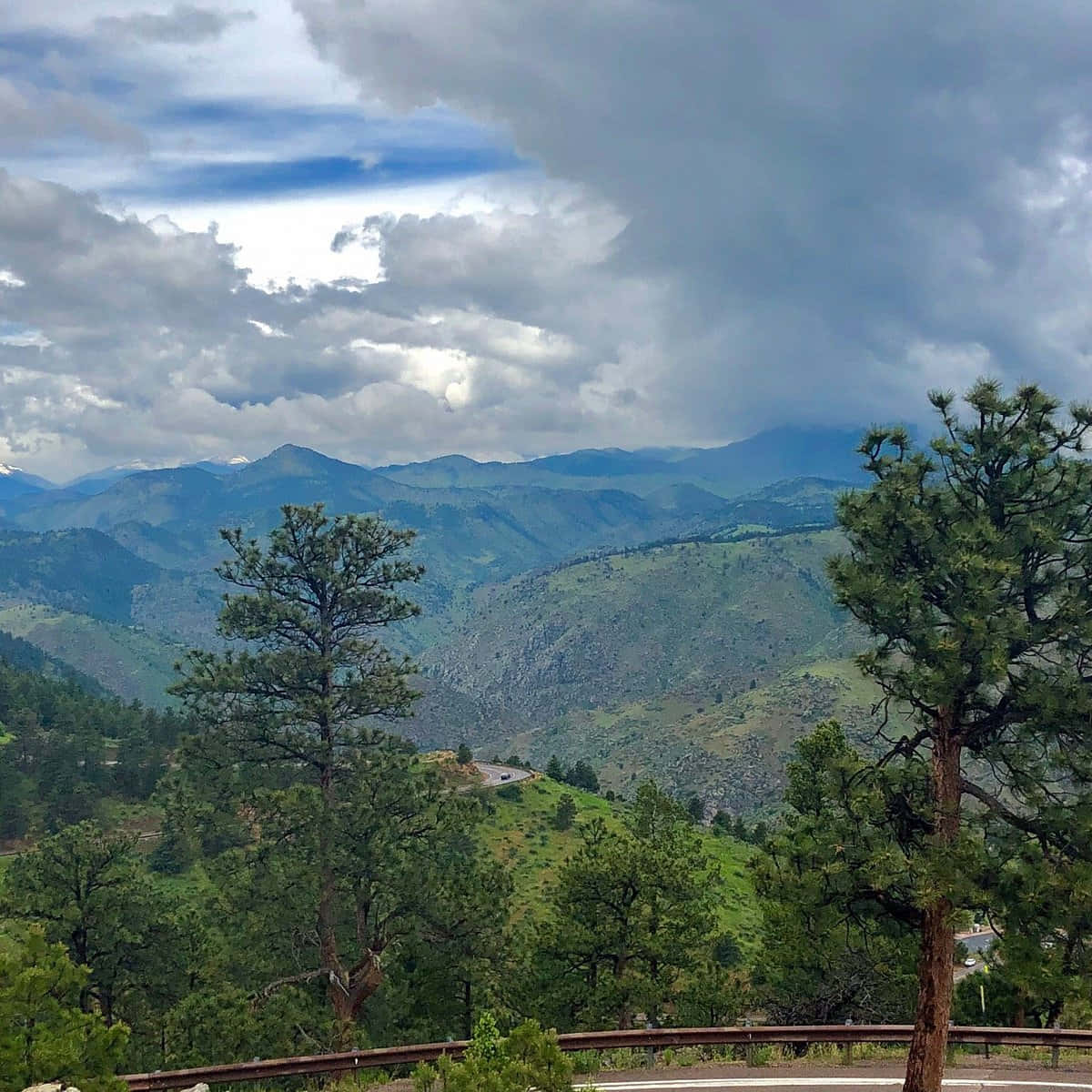 Colorado Mountain 桌布 壁紙 — 一條蜿蜒的山路穿過鬱鬱蔥蔥的綠色山谷，在戲劇性的多雲天空下，遠處有白雪覆蓋的山峰。