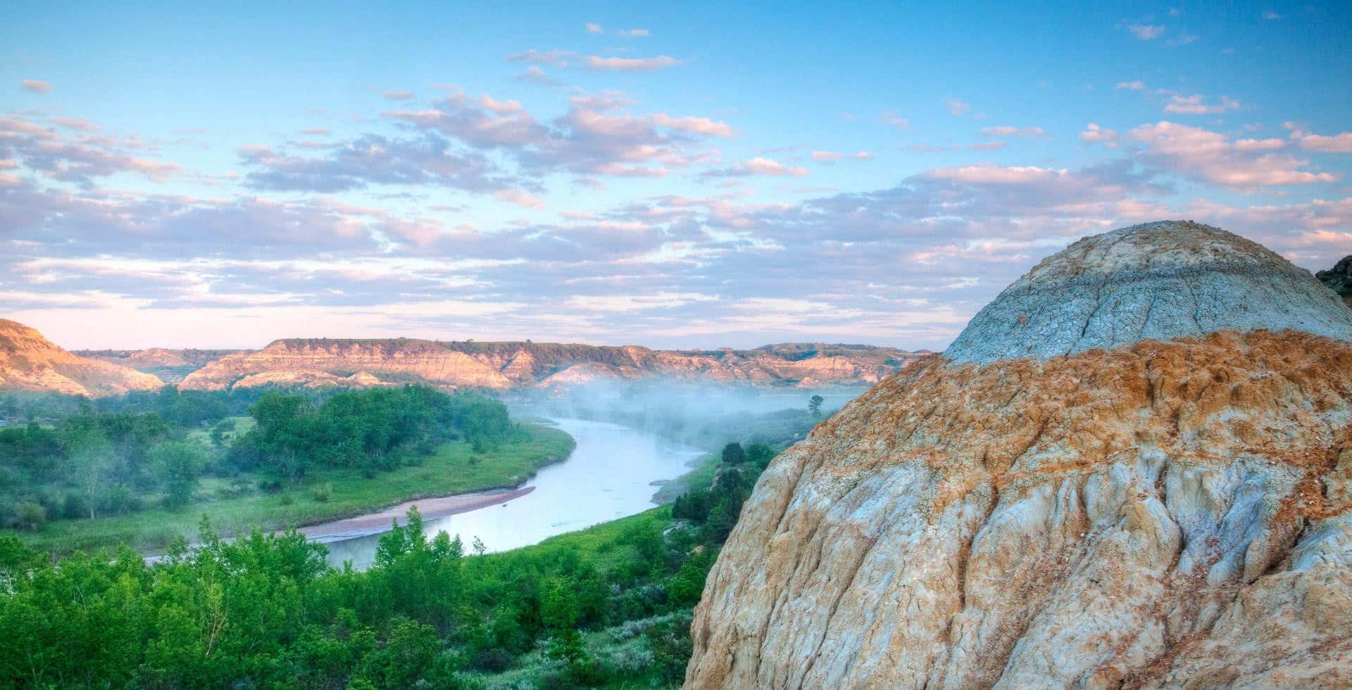 North Dakota 桌布 壁紙 — 一條蜿蜒的河流在日出時流經霧氣瀰漫的峽谷，層層疊疊的岩石形態和茂密的綠植在柔和粉彩的天空下。