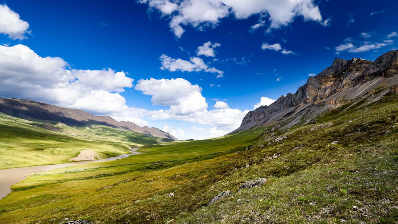 720p Nature 桌布 壁紙 — 一條蜿蜒的河流流經充滿活力的綠色山谷，兩側是崎嶇的山脈，上方是明亮的藍天和蓬鬆的白雲。