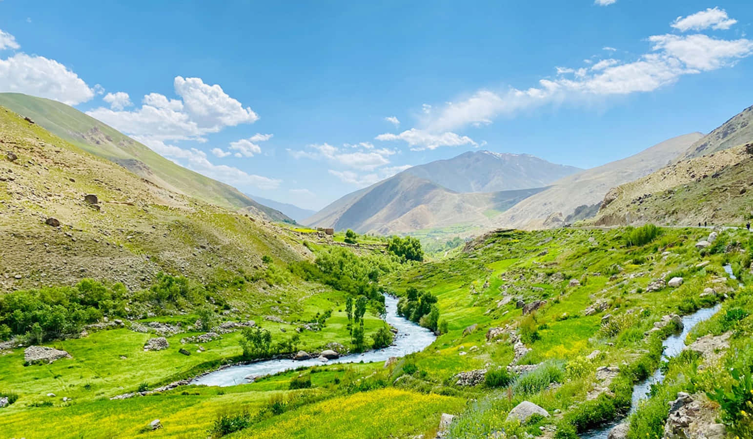 Afghanistan 桌布 壁紙 — 一條蜿蜒的河流流經充滿活力的綠色山谷，兩旁是陽光照耀的山脈，天空湛藍，雲朵點綴。