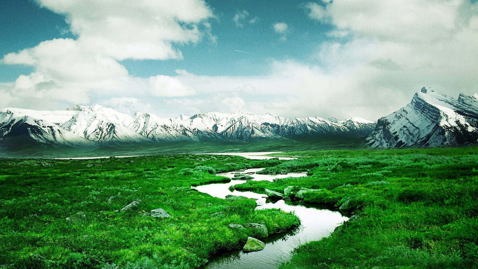1920X1080 桌布 壁紙 — 一條蜿蜒的河流流經充滿活力的綠色草地，白雪覆蓋的山脈在部分多雲的天空下聳立。