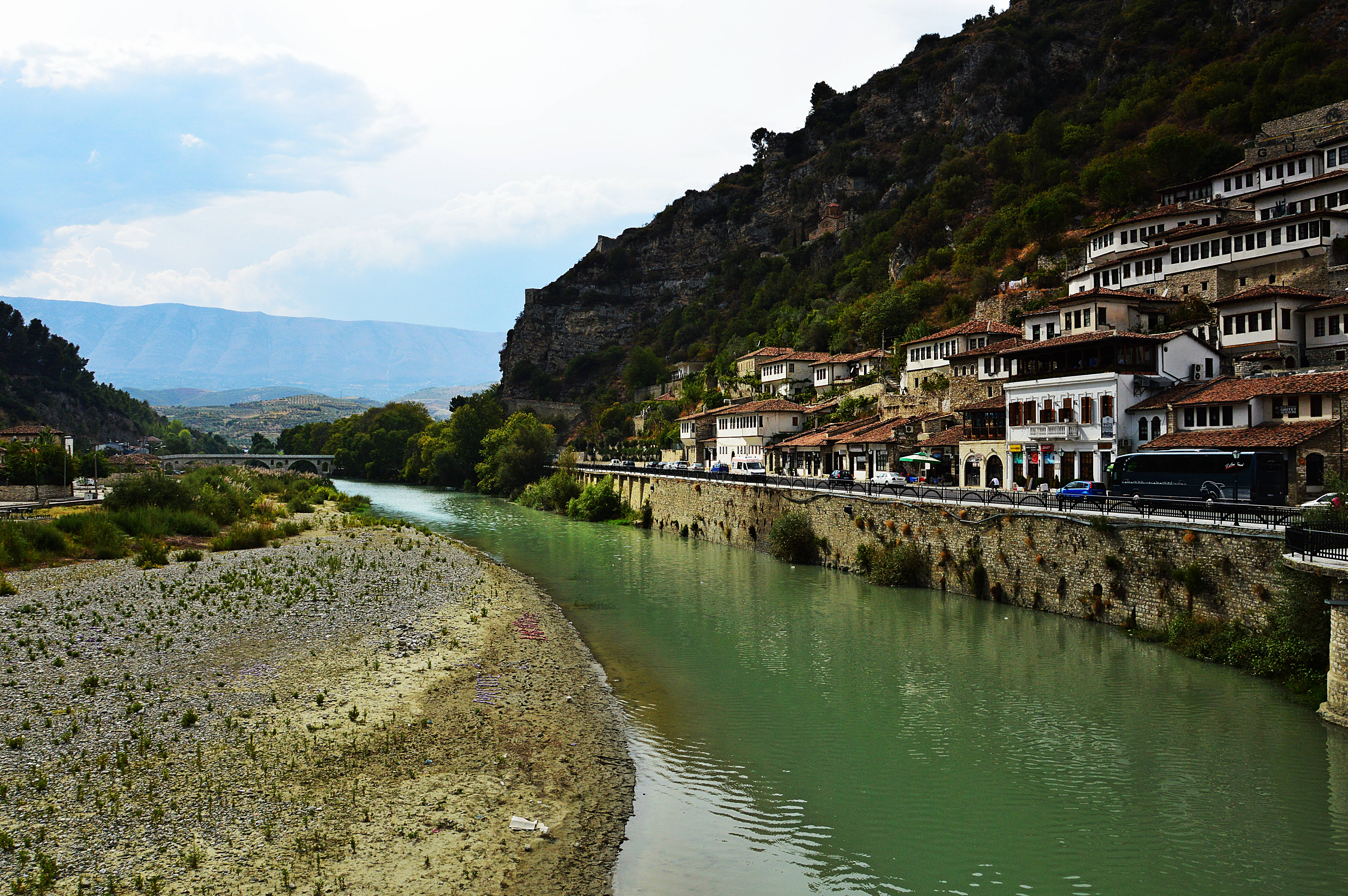 Albania 桌布 壁紙 — 一條蜿蜒的河流流過一個山坡村莊，村莊裡有陶土屋頂的房子，天空部分多雲。