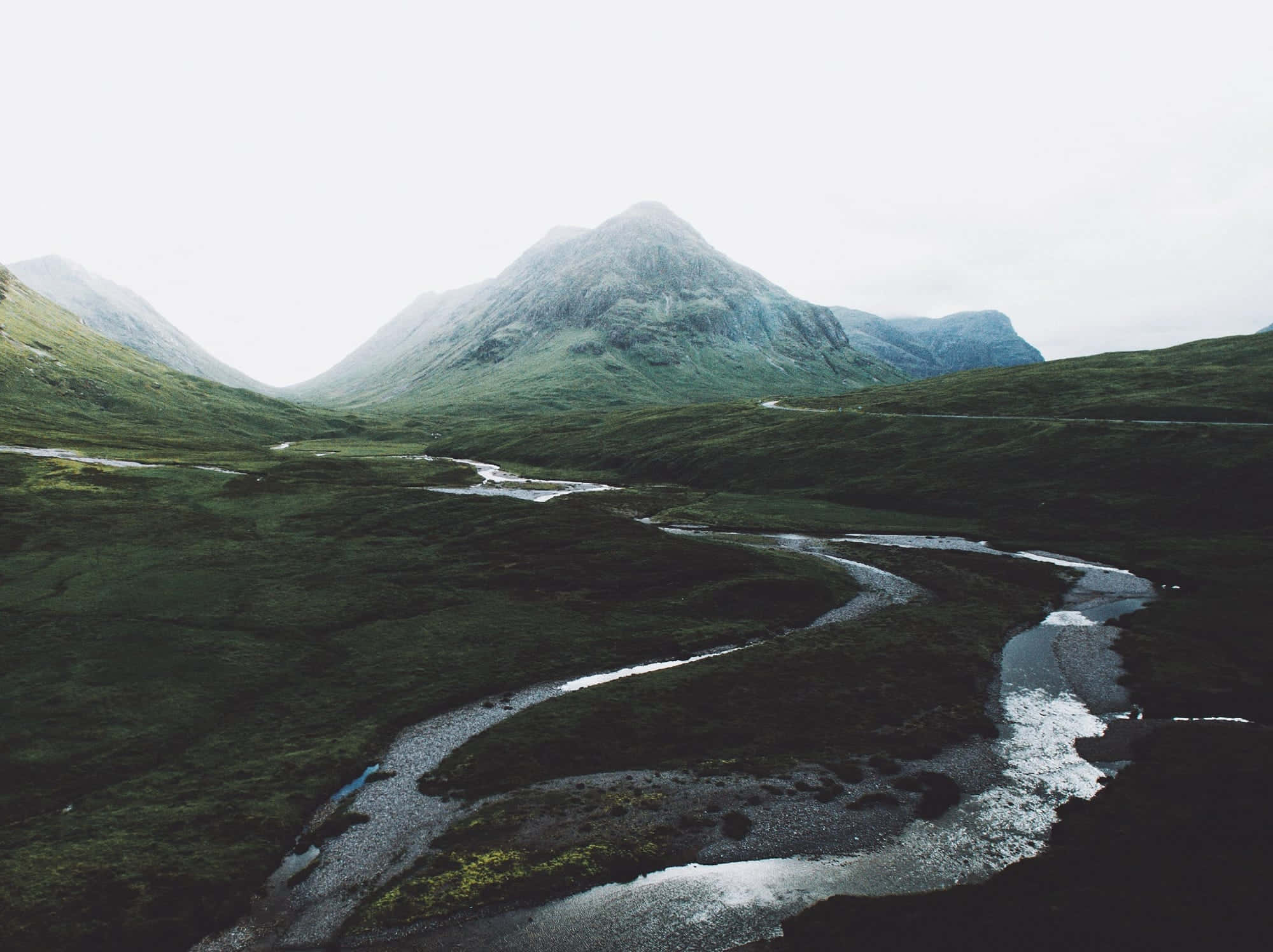 Vsco Laptop 桌布 壁紙 — 一條蜿蜒的河流穿過一個鬱鬱蔥蔥的綠色山谷，兩旁是高聳的霧濛濛的山脈，天空陰沉。