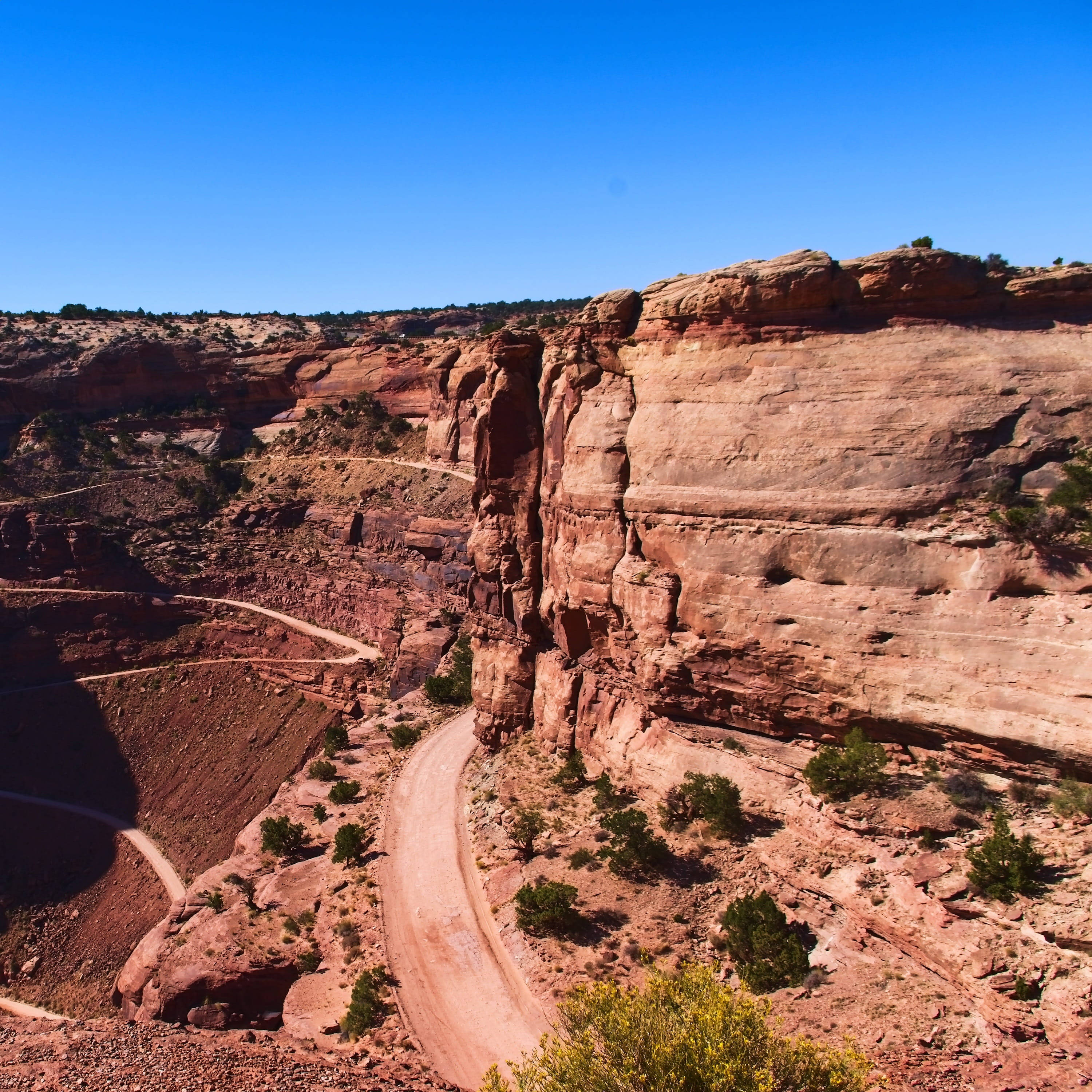 Canyonlands National Park 桌布 壁紙 — 一條蜿蜒的泥土路在沙漠峽谷中，穿過高聳的紅色岩層，在清澈的藍天下蜿蜒而行。