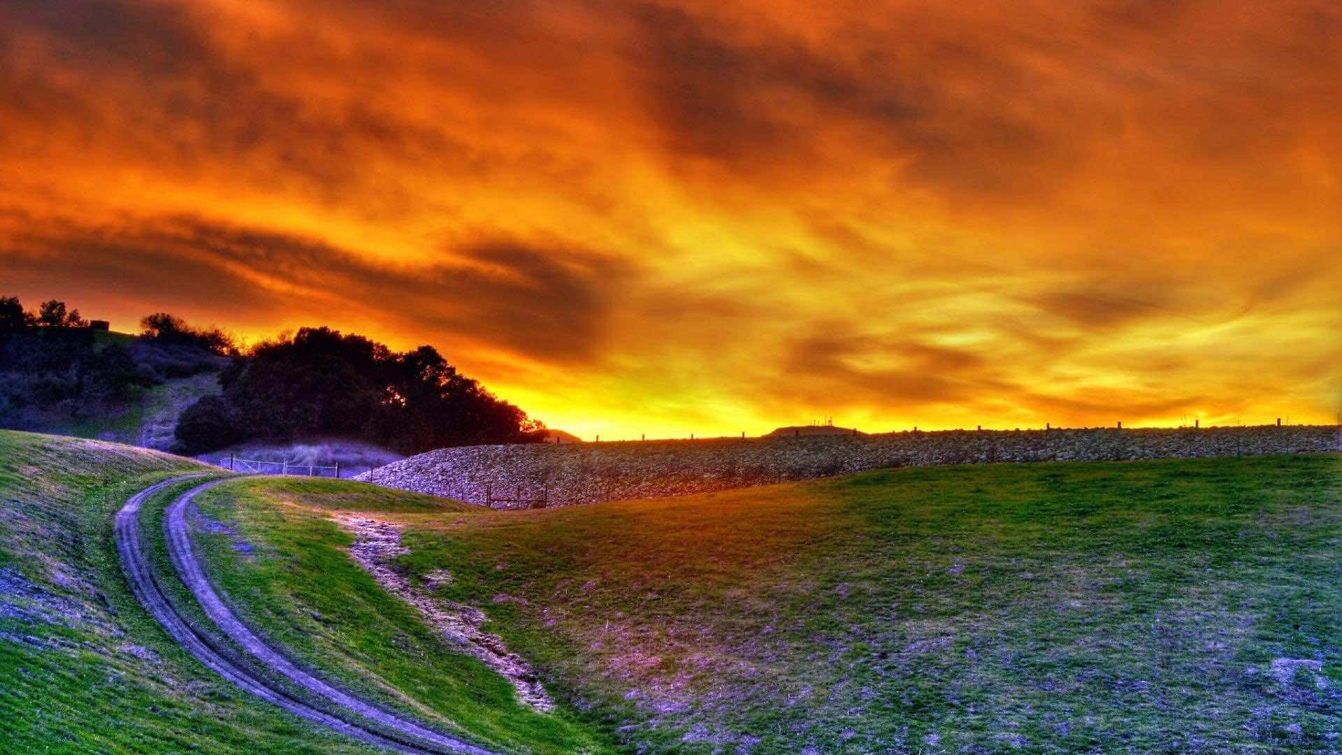 Beautiful Landscape 桌布 壁紙 — 一條蜿蜒的泥土路在火紅橙黃色的日落天空下，穿過充滿活力的綠色山丘。