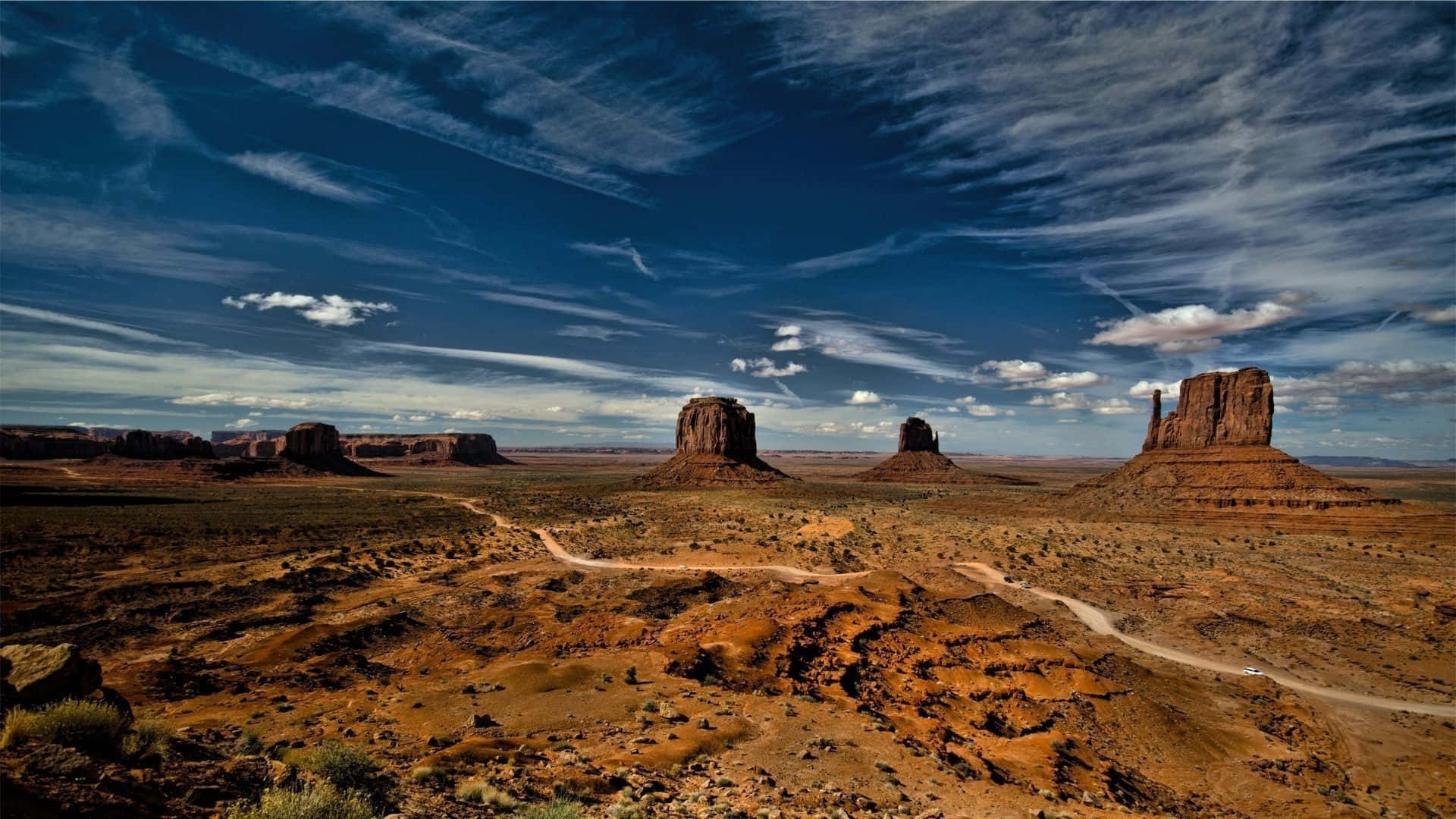 Arizona 桌布 壁紙 — 一條蜿蜒的泥土路穿過廣闊的沙漠景觀，點綴著高聳的紅色岩層，上方是戲劇性的藍天和縷縷白雲。