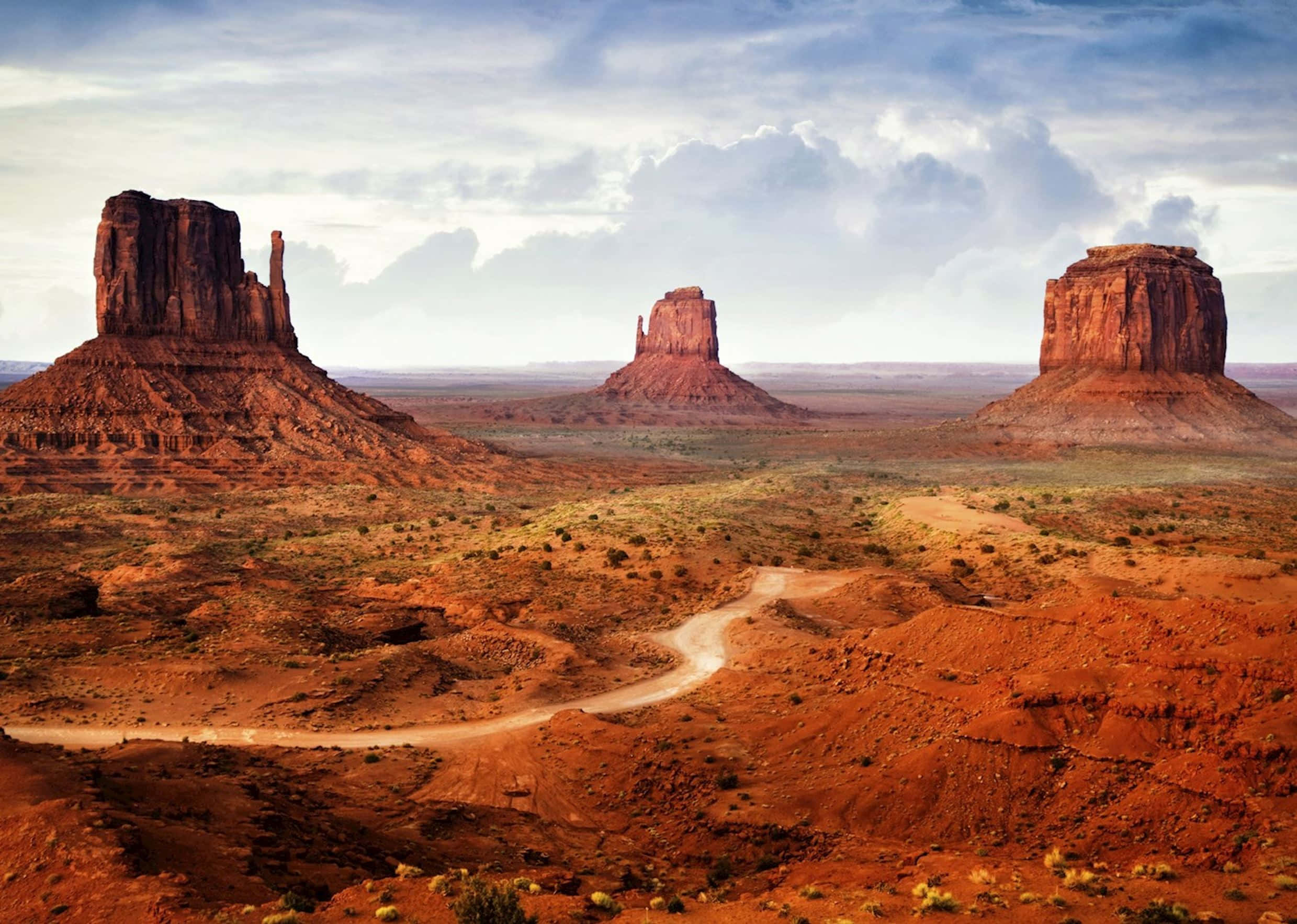 Arizona 桌布 壁紙 — 一條蜿蜒的泥土路穿過廣闊的紅色沙漠景觀，點綴著高聳的砂岩但丁，天空多雲。