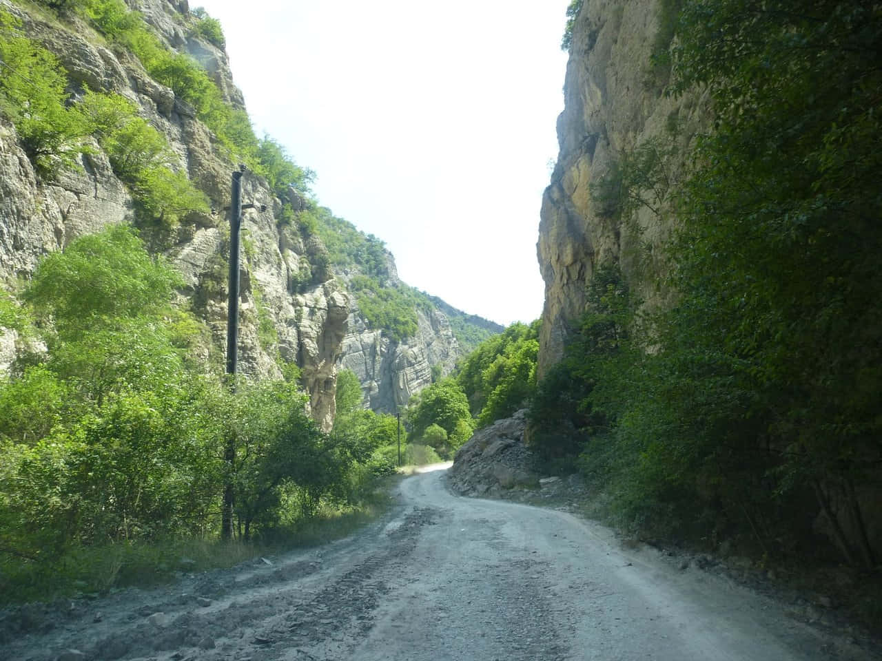 Road 桌布 壁紙 — 一條蜿蜒的泥土路穿過狹窄的峽谷，兩旁是陡峭的岩石峭壁，在明亮的日光下覆蓋著茂密的綠色植被。