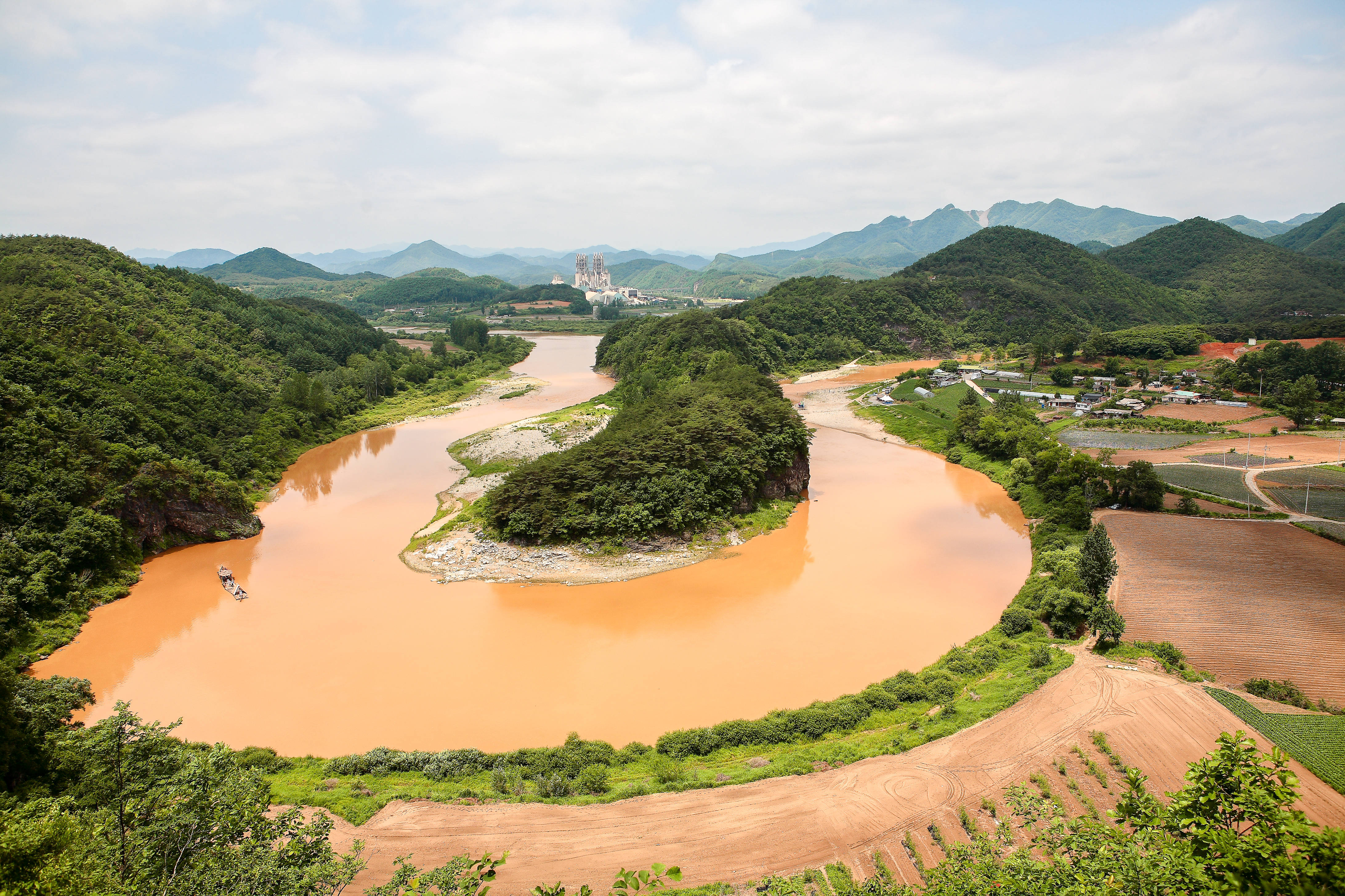 Topography 桌布 壁紙 — 一條蜿蜒的泥濘河流穿過鬱鬱蔥蔥的綠色山丘和農田，天空多雲，遠處有工廠。