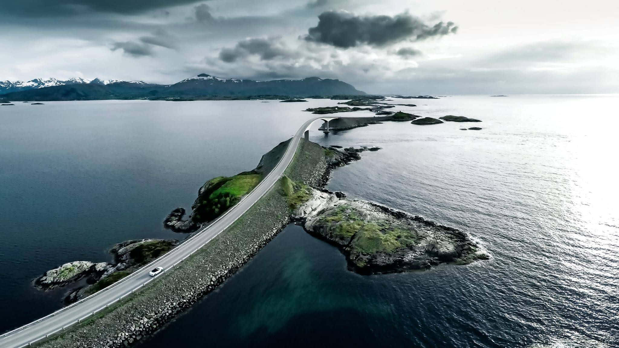 Road 桌布 壁紙 — 一條蜿蜒的海岸公路在戲劇性的多雲天空下，延伸穿過岩石小島和深藍色水域。