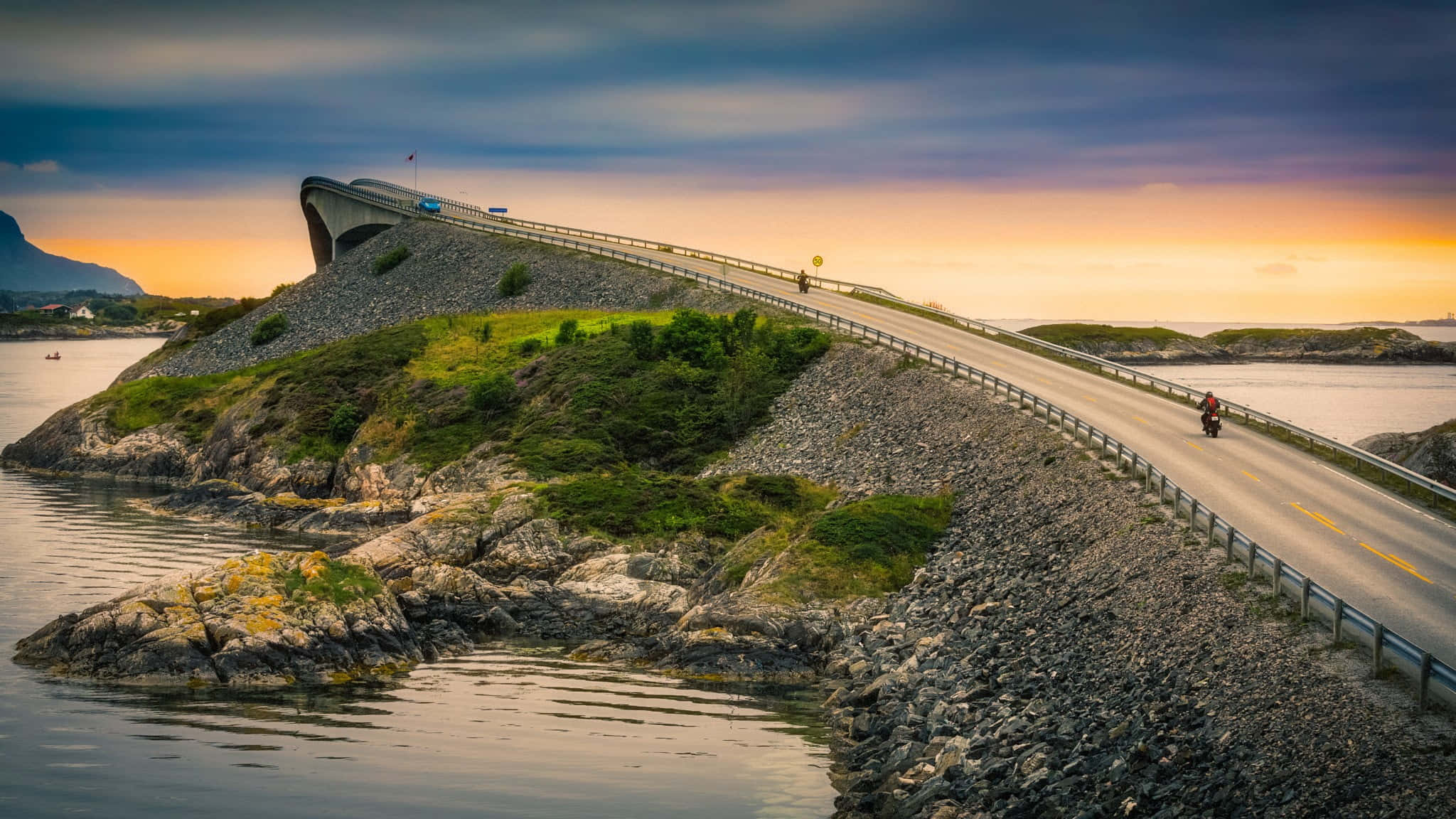 Road 桌布 壁紙 — 一條蜿蜒的海岸公路在日落時分戲劇性地彎曲穿過岩石島嶼，一名孤獨的摩托車騎士騎向地平線。