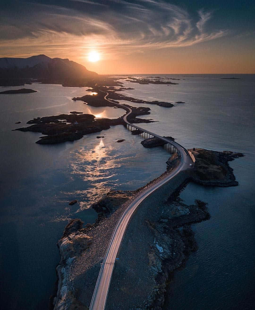 Road 桌布 壁紙 — 一條蜿蜒的海岸公路在日落時分穿過岩石島嶼，金色的光線映照在平靜的水面上。