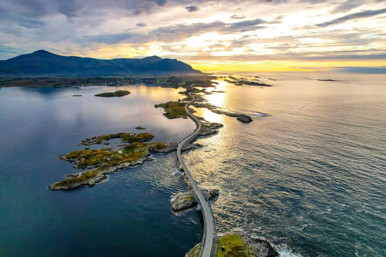Road 桌布 壁紙 — 一條蜿蜒的海岸公路在日落時分穿過岩石島嶼，金色的光線反射在海洋和遠處的山脈上。